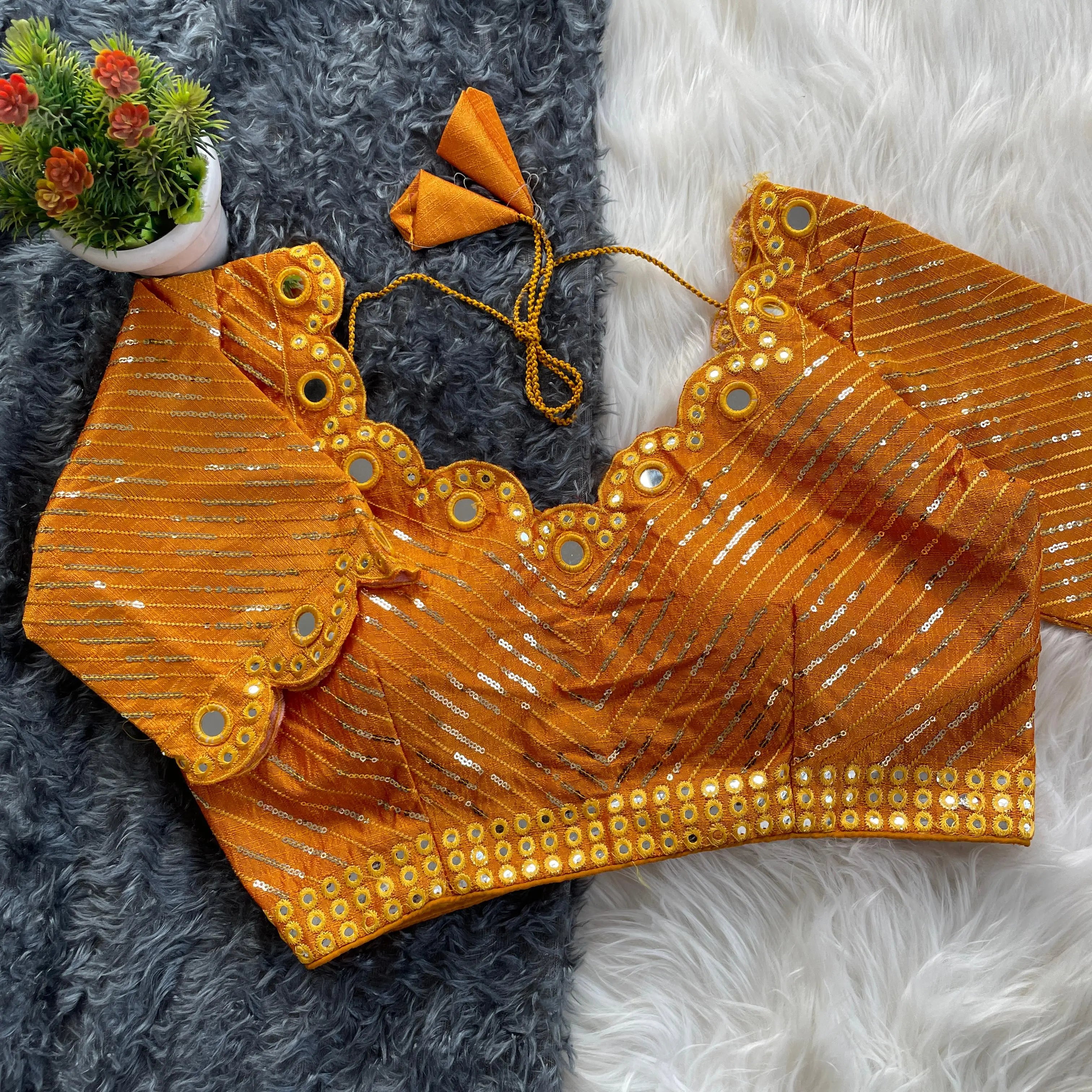Orange bridal blouse with scallop sleeves, featuring circular mirror work and silver sequin stripes, decorative borders on sleeves and hem, and tie-up tassels, displayed on a textured grey and white fur background with a small potted plant nearby.
