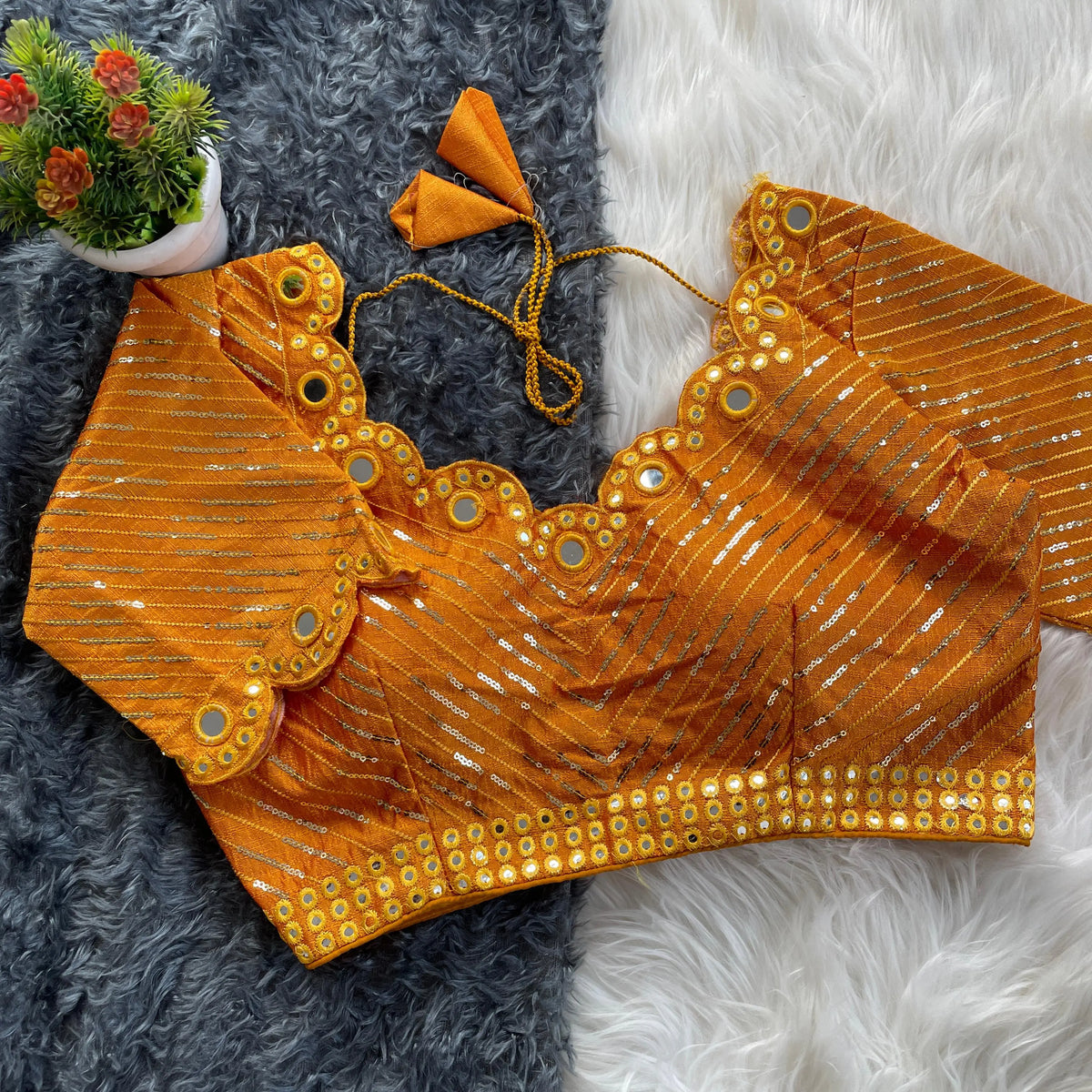 Orange bridal blouse with scallop sleeves, featuring circular mirror work and silver sequin stripes, decorative borders on sleeves and hem, and tie-up tassels, displayed on a textured grey and white fur background with a small potted plant nearby.