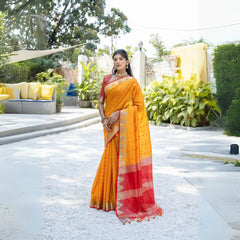 A woman models a vibrant Orange Tussar Silk Saree with scattered dark woven motifs. The saree has a gold border and a pallu featuring wide bands of rich red and gold weaving. She wears a red blouse and gold Kundan jewelry, posing outdoors.