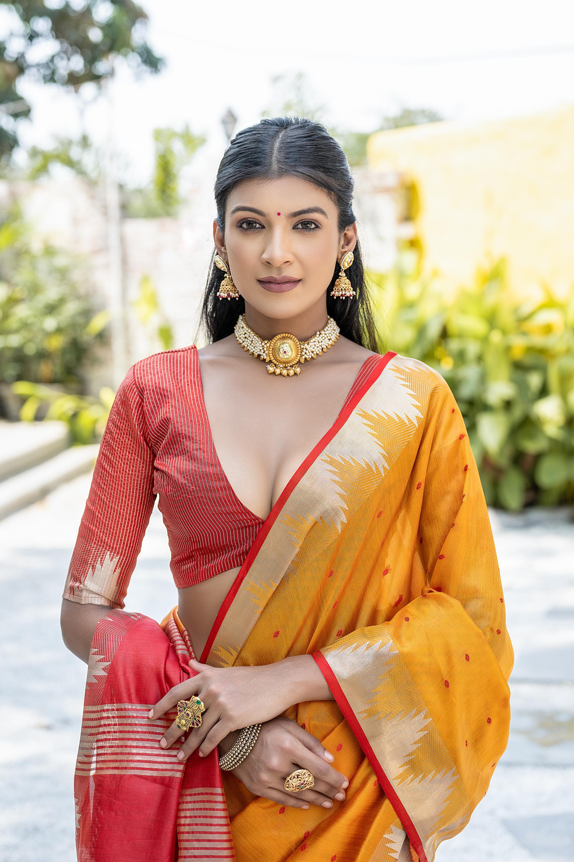A woman models a vibrant Orange Tussar Silk Saree with scattered dark woven motifs. The saree has a gold border and a pallu featuring wide bands of rich red and gold weaving. She wears a red blouse and gold Kundan jewelry, posing outdoors.