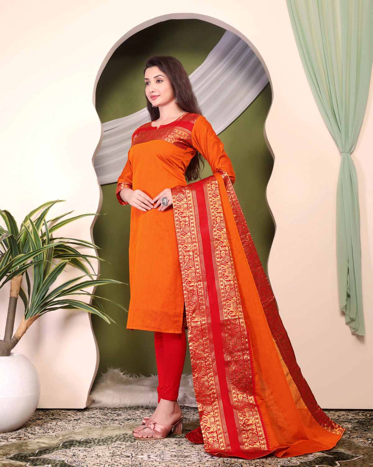 Woman wearing an orange and red traditional outfit with a decorative border, standing in a room with a plant and curtain.