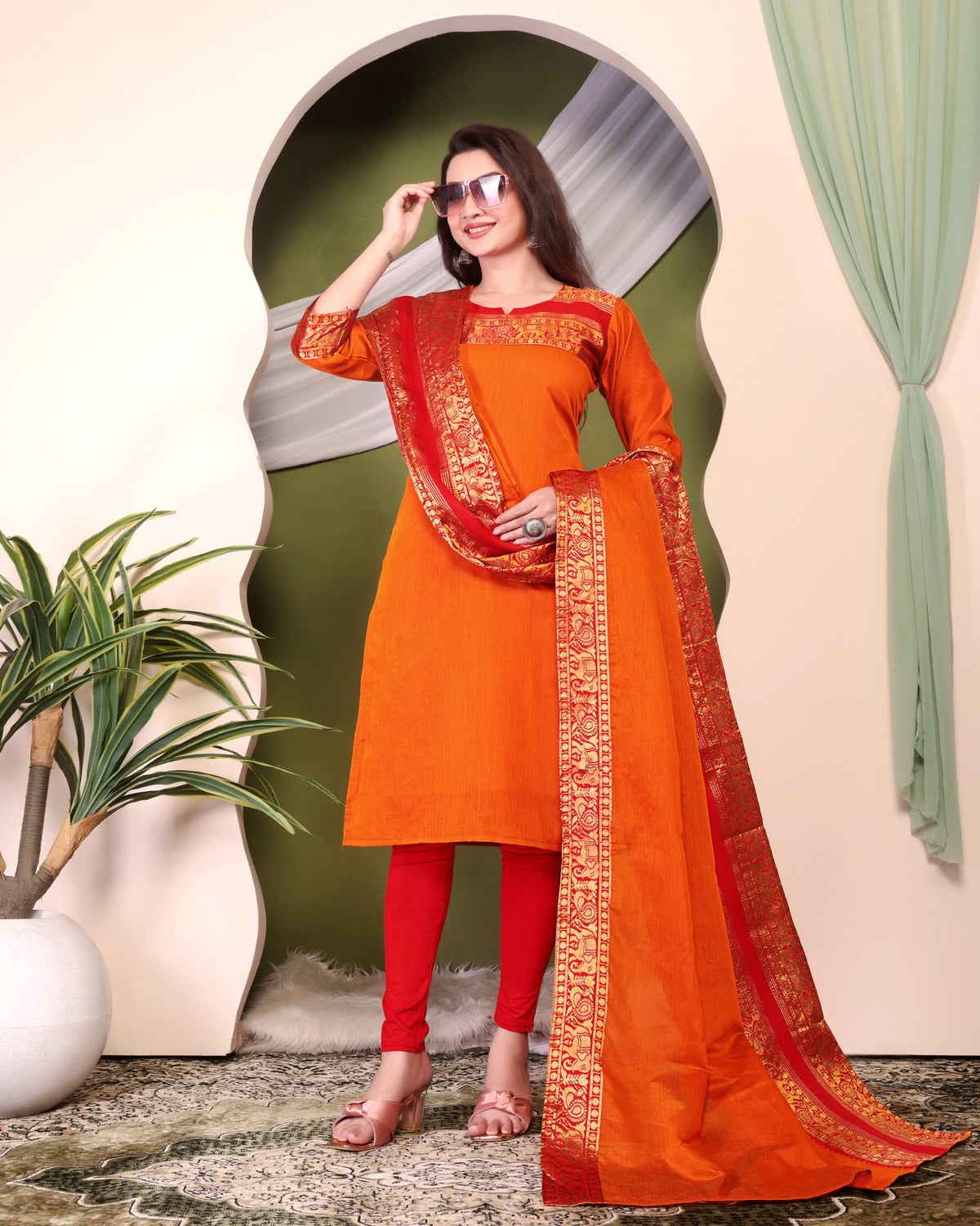 Woman wearing an orange and red traditional outfit with a green curtain and plant in the background