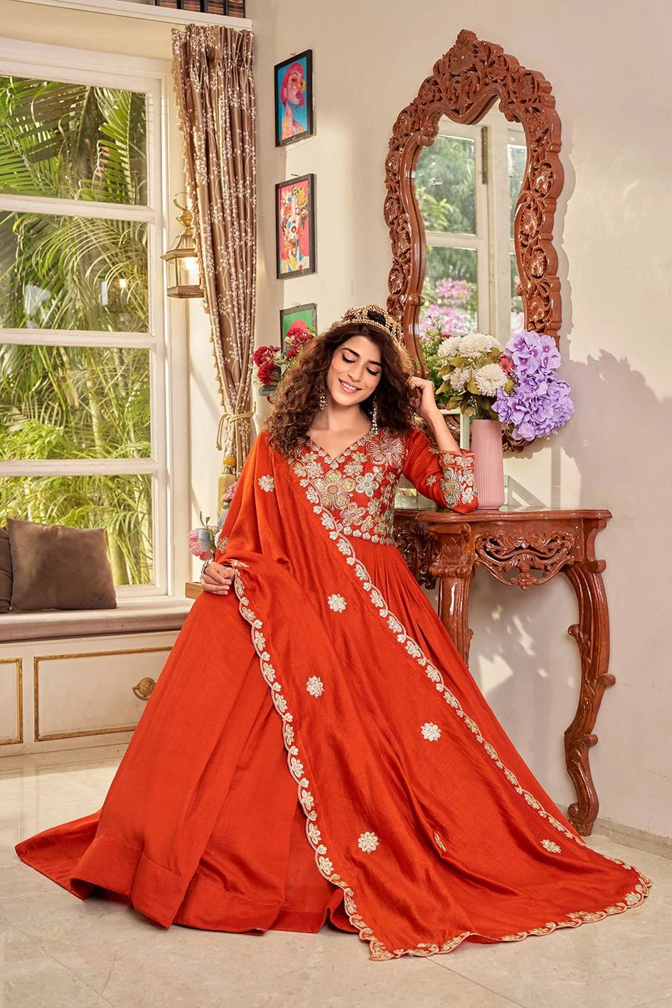 Woman in a red saree with gold details standing in a room with a decorative mirror and floral arrangement.