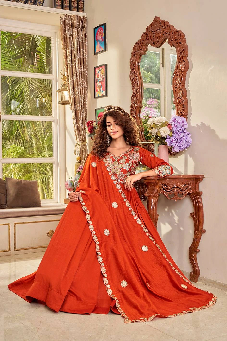 Woman in a red saree with gold details standing in a room with a decorative mirror and window.
