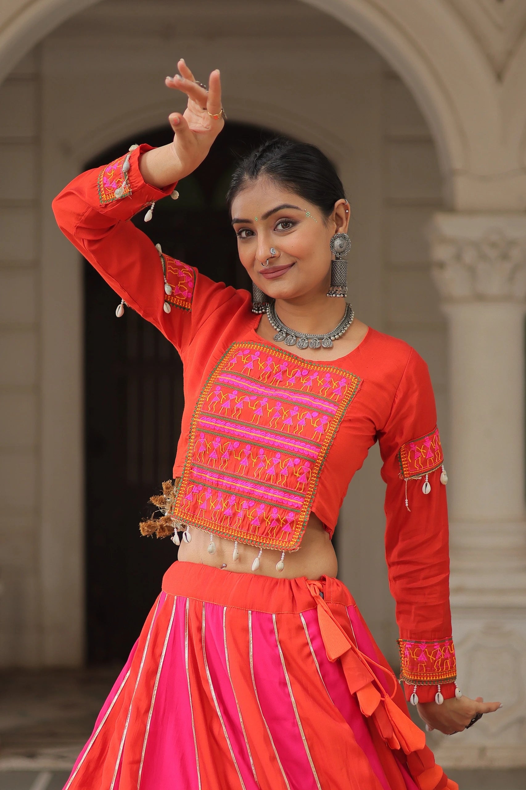 Woman in traditional red and pink outfit with intricate designs, standing in an archway.