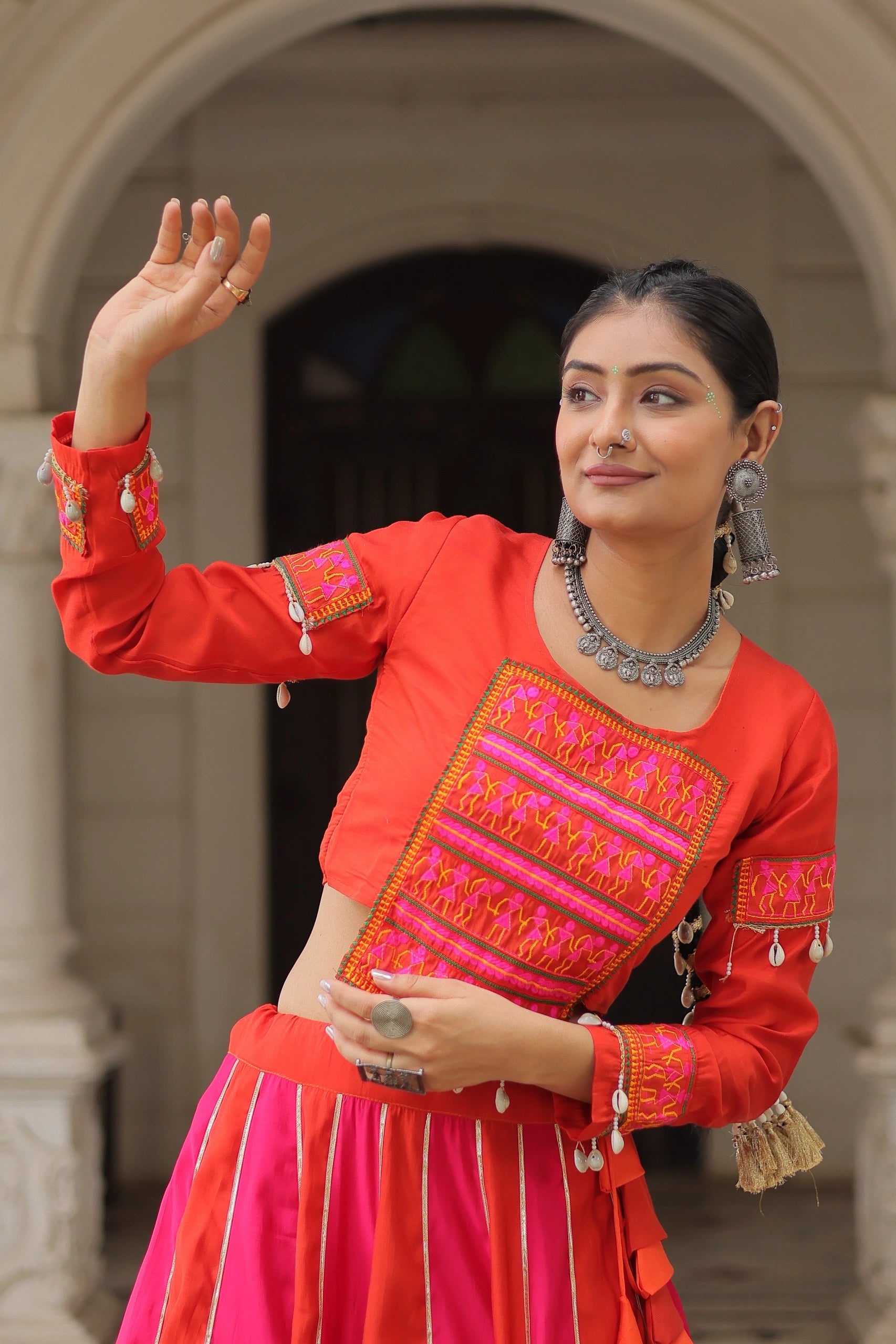 Woman in traditional red and pink outfit with intricate patterns, standing in an archway.