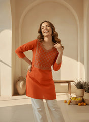 Woman wearing an orange traditional outfit in a sunlit room with plants and fruit.