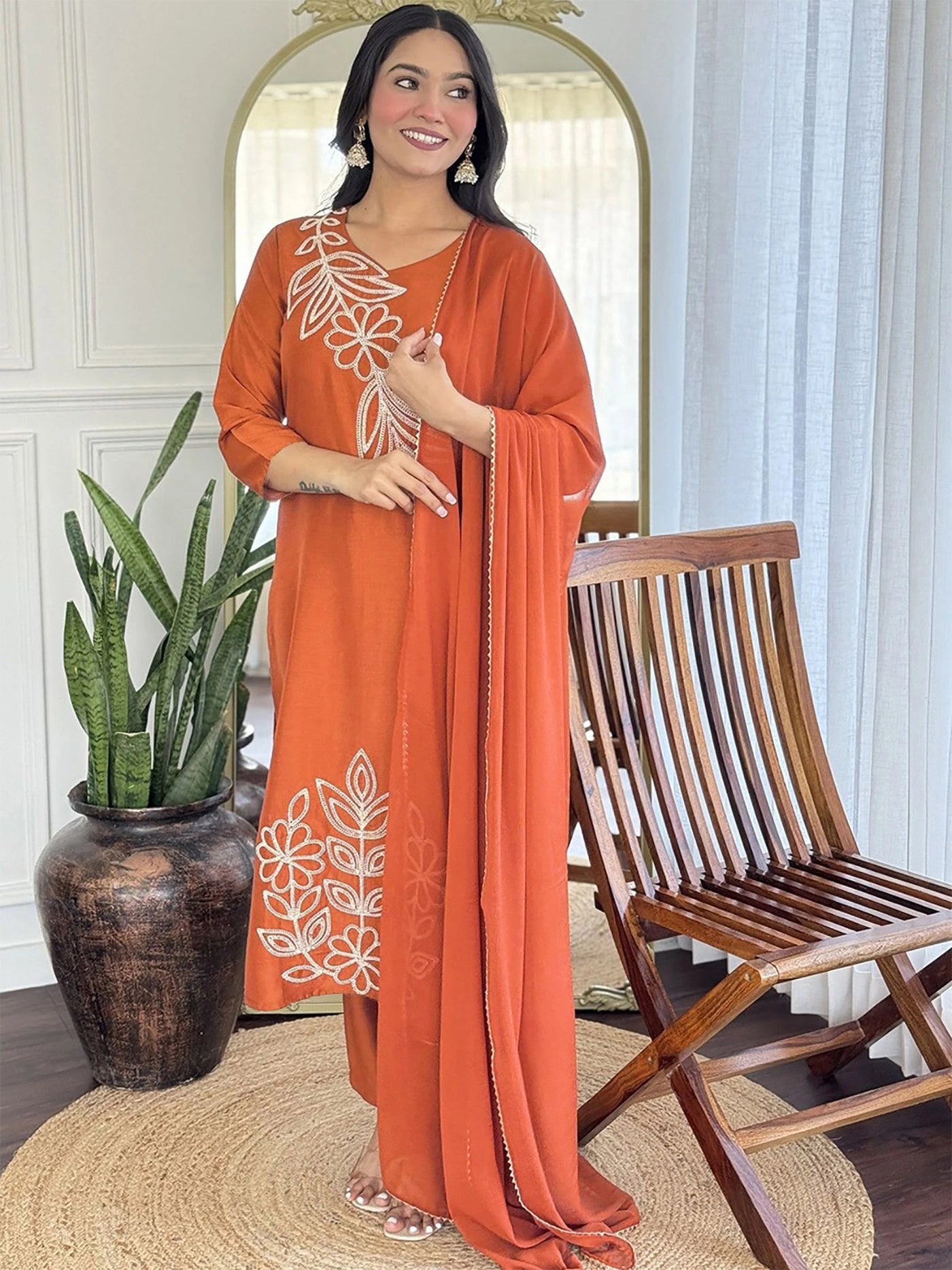 Woman in an orange embroidered outfit standing in a room with a wooden chair and plant.