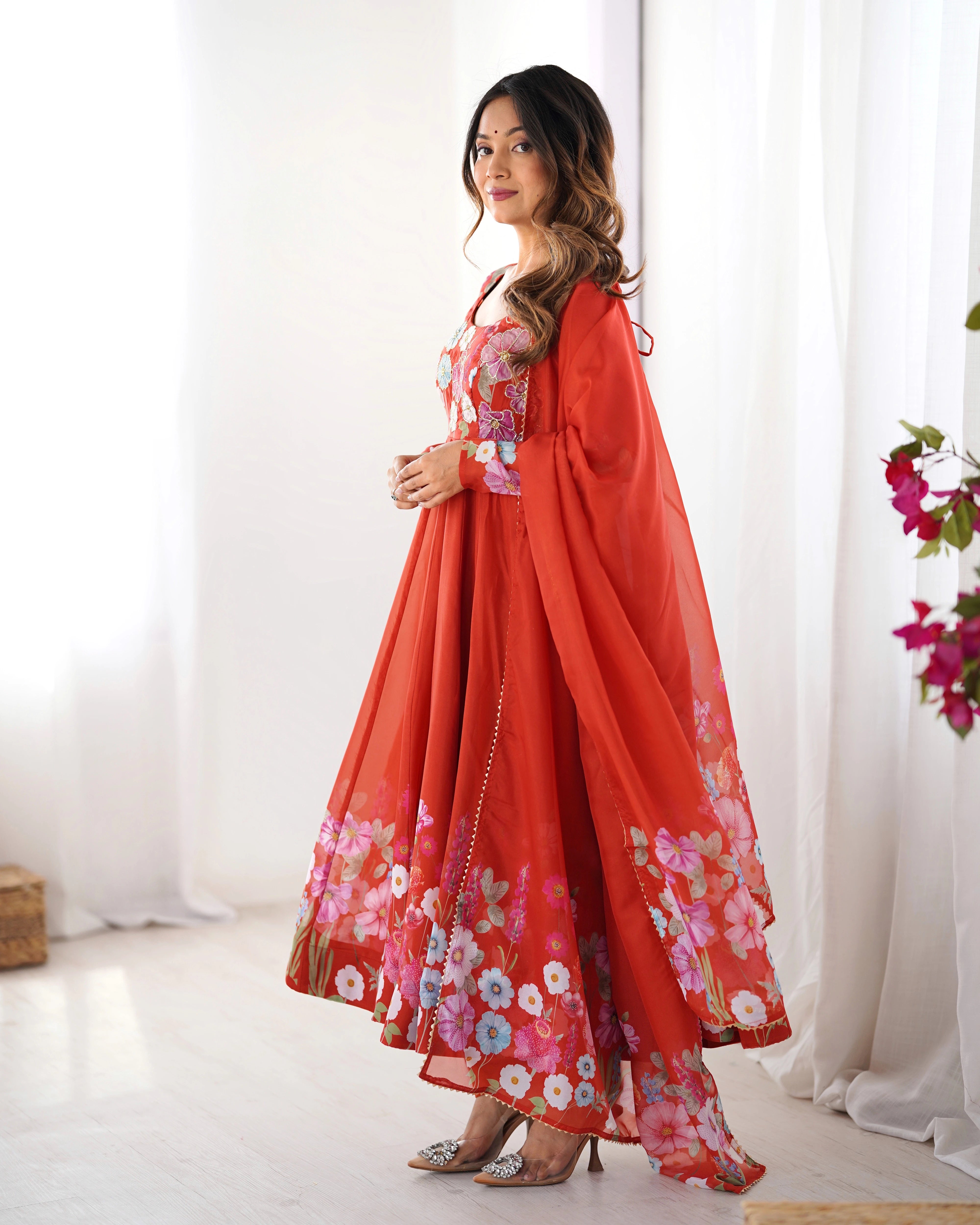 Woman wearing a red floral dress standing in a bright room with white curtains.