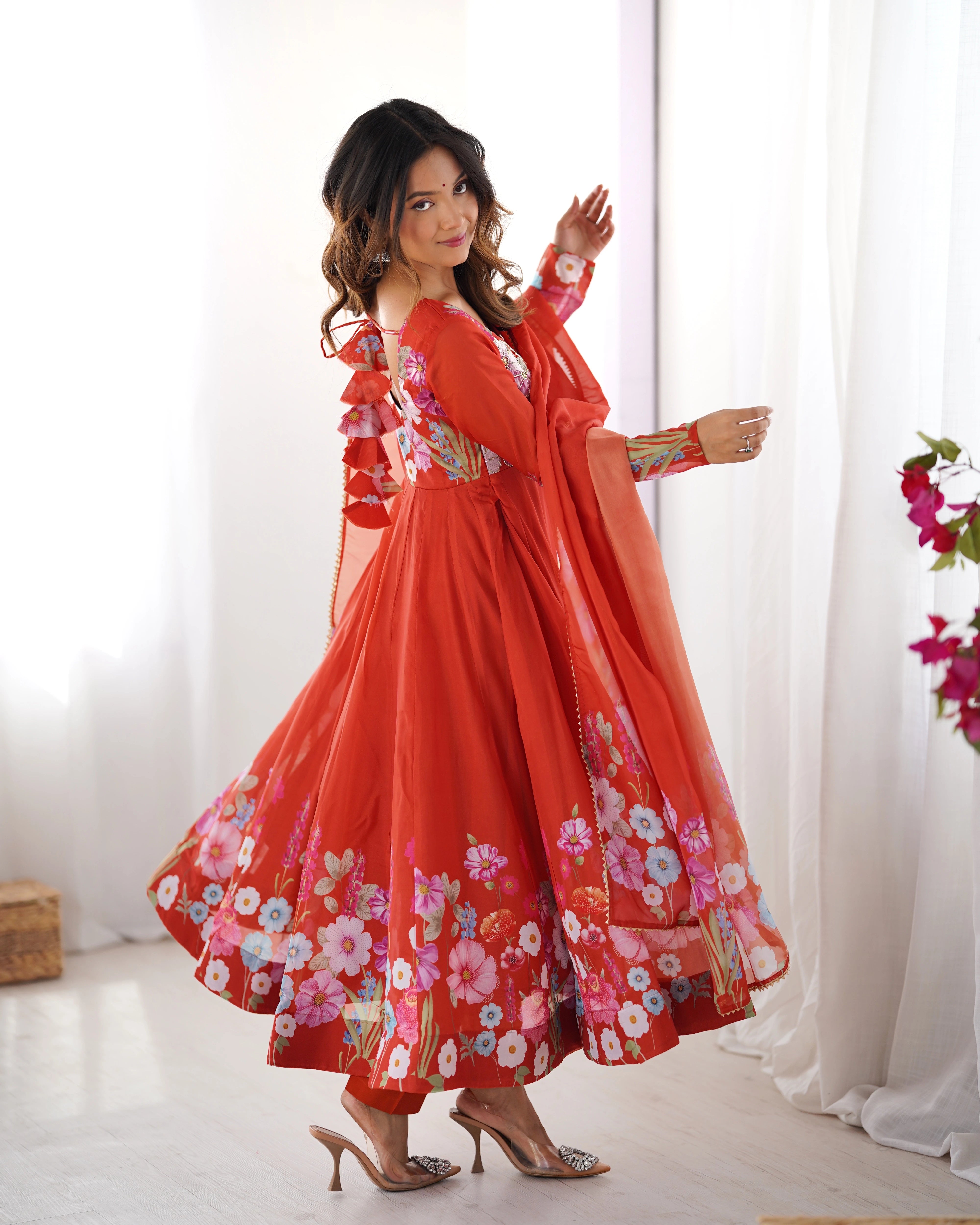 Woman wearing a red floral dress standing in a bright room with white curtains.