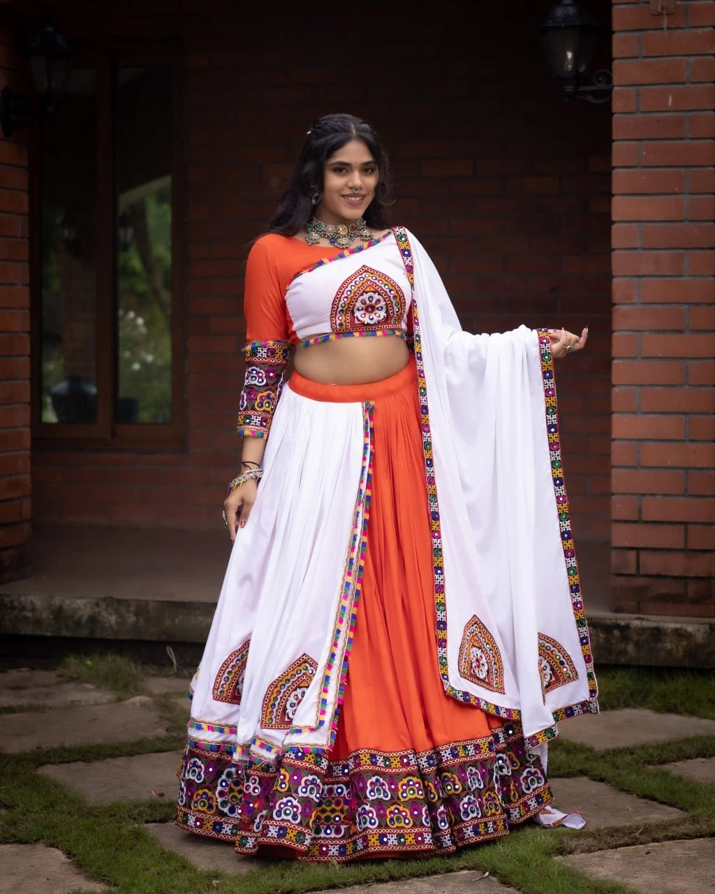 Woman in traditional orange and white outfit with colorful patterns standing outdoors.