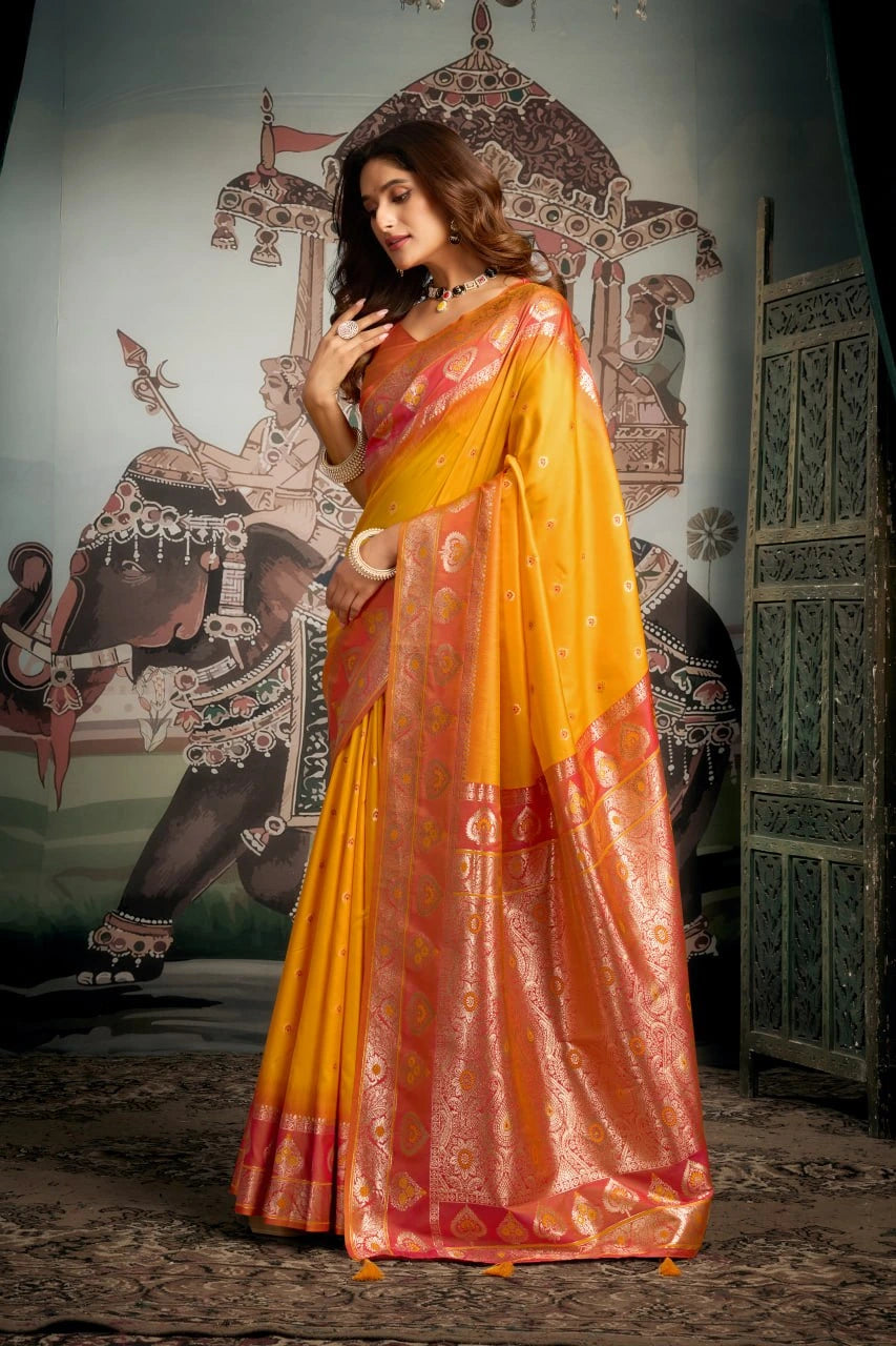 Woman in a yellow and orange saree standing in front of a decorative wall.
