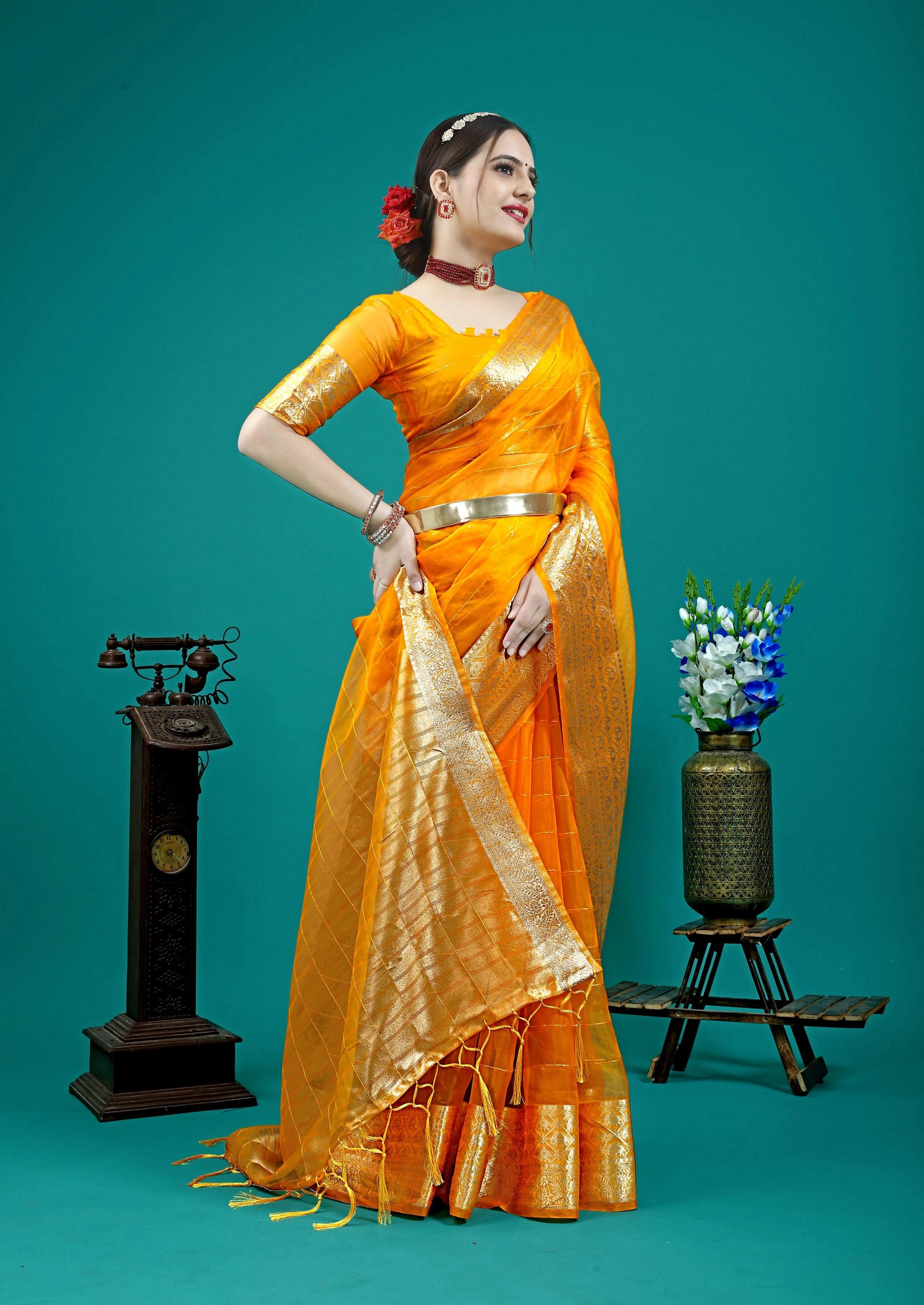 Woman in an orange saree standing against a teal background with decorative elements.