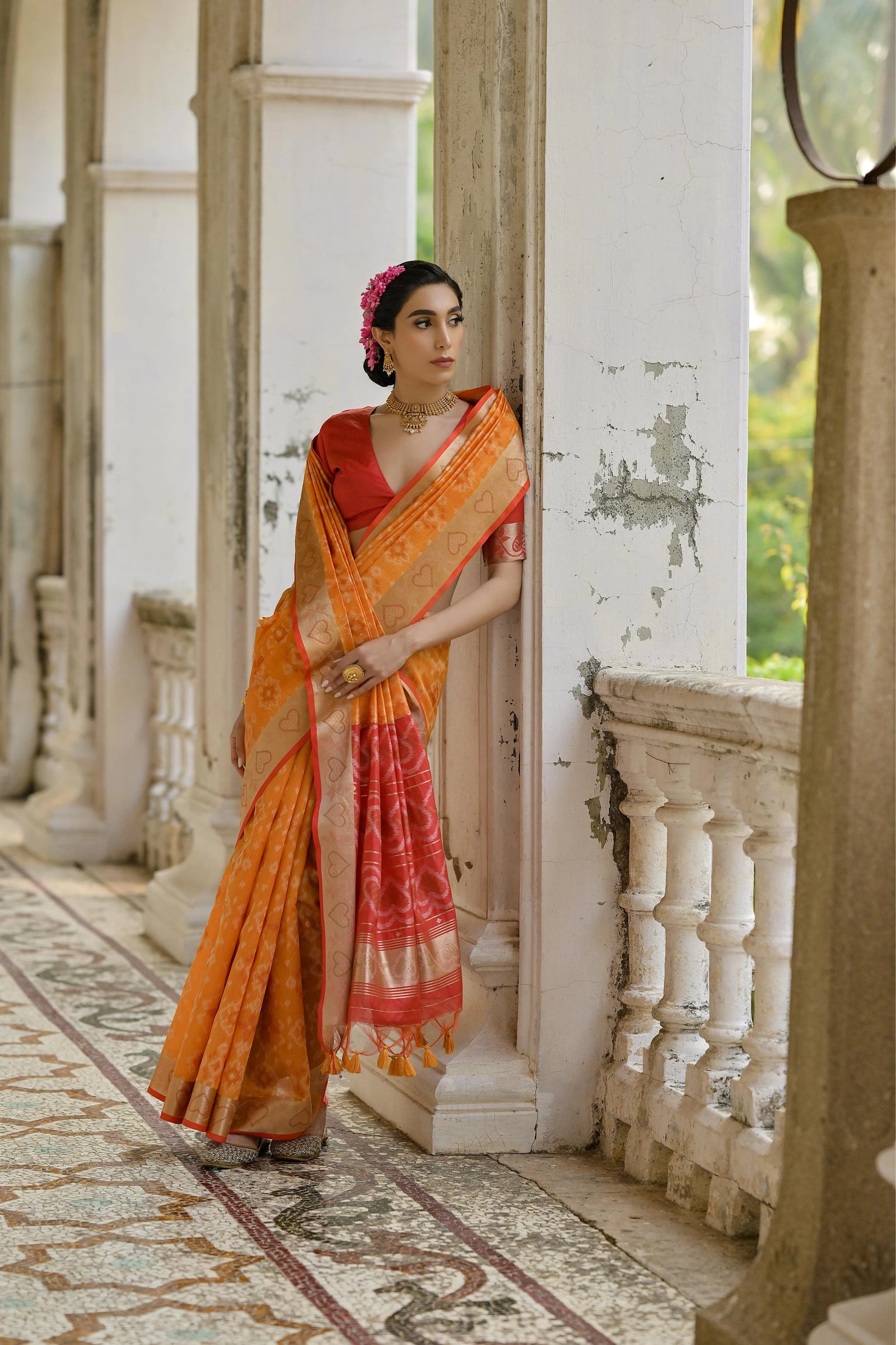 Woman in an orange and red saree standing in a decorative interior setting.