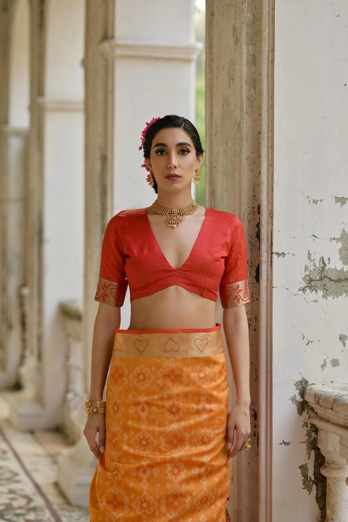 Woman wearing a red blouse and orange skirt standing in an outdoor setting with architectural elements.