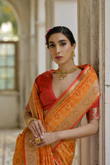 Woman in an orange saree with a red blouse, standing indoors.