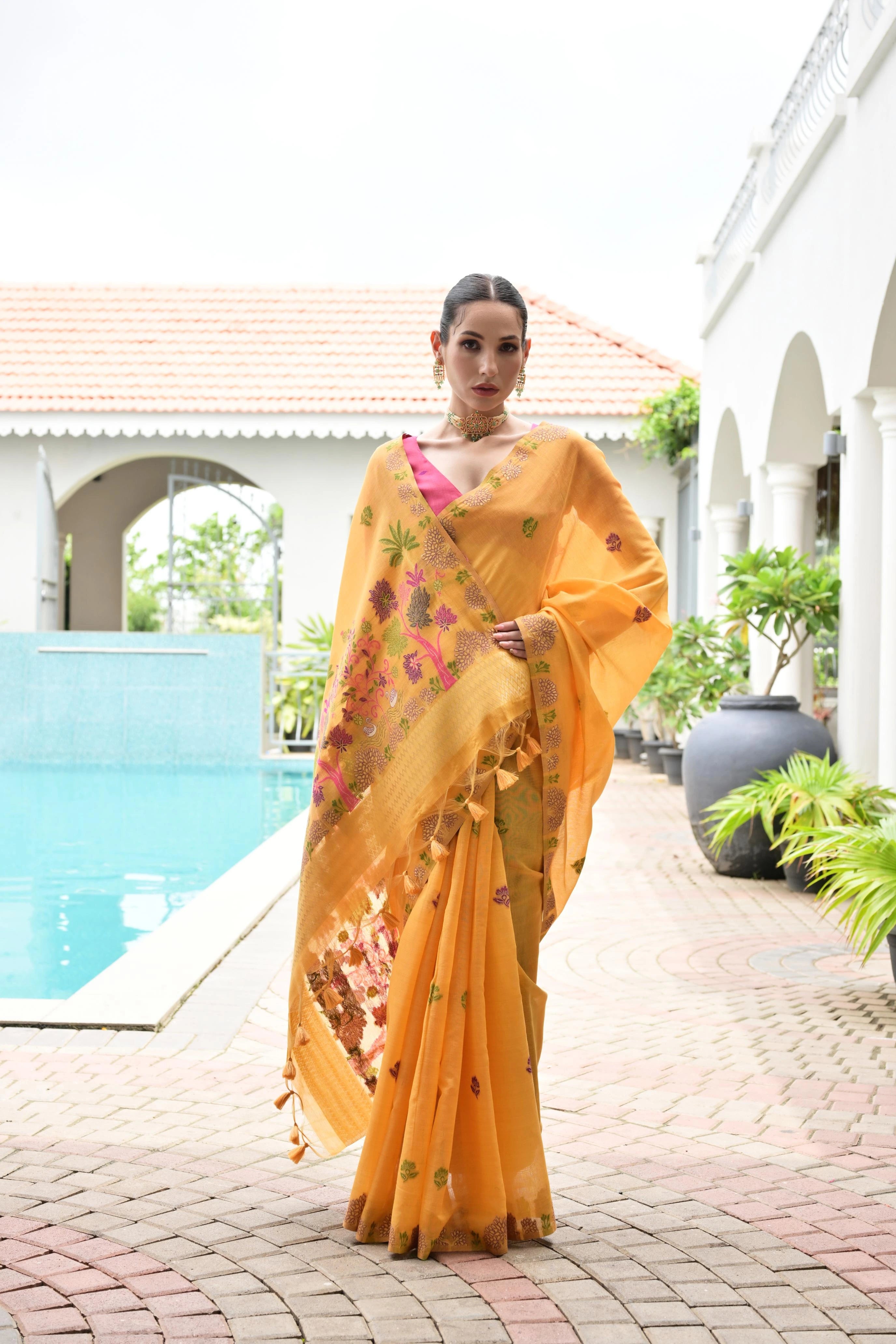 Woman in a yellow saree with a floral pattern standing by a pool.