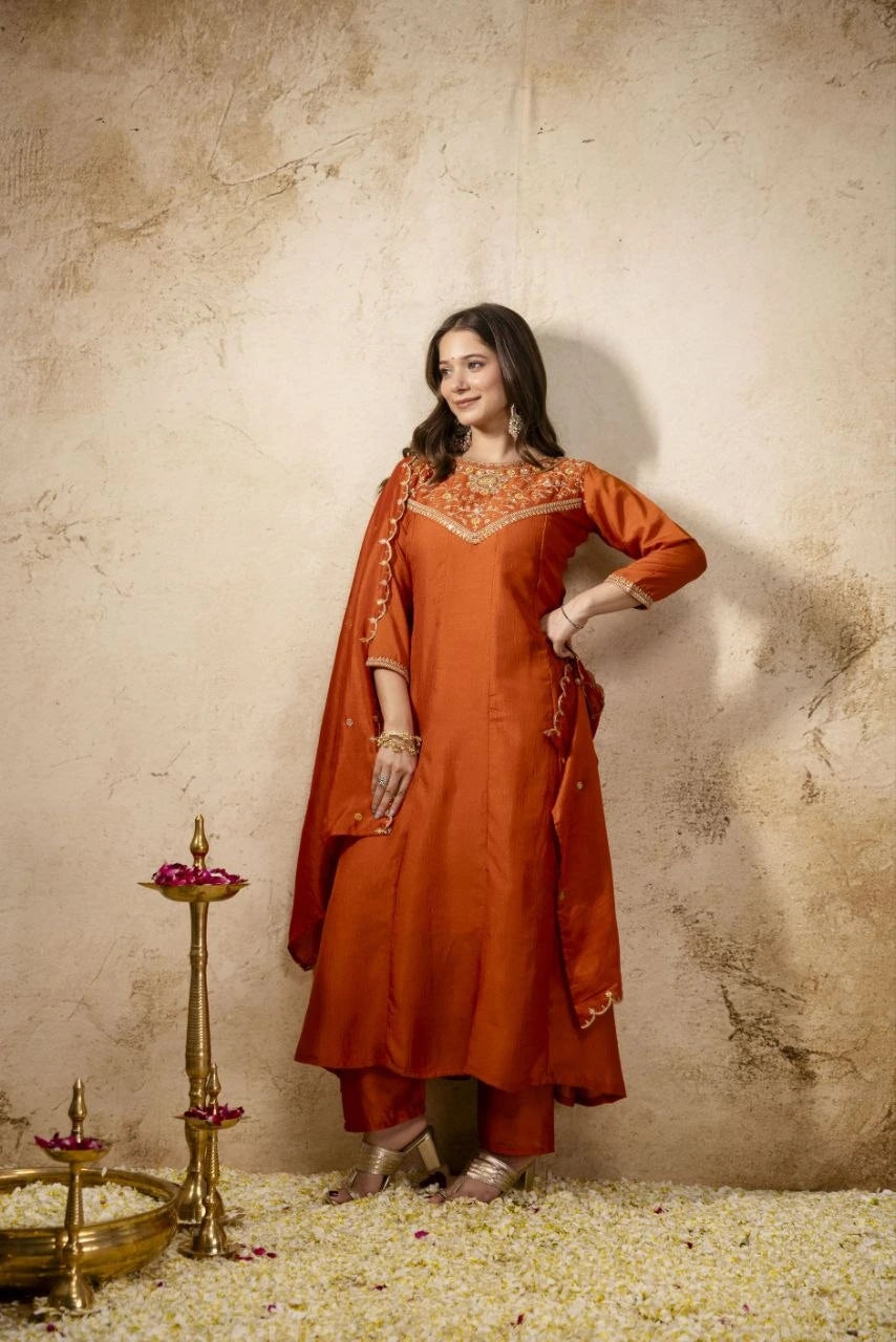 Woman in an orange traditional outfit with embroidery, standing against a textured beige wall.