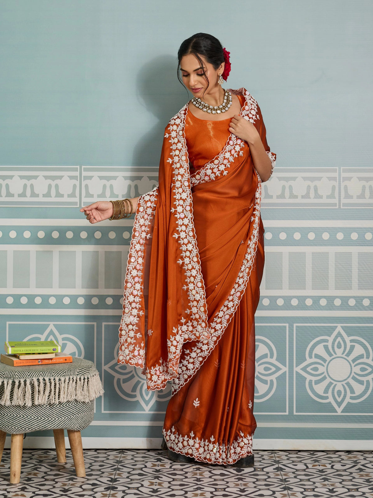 Woman in an orange saree with white patterns standing in a room with decorative wallpaper.