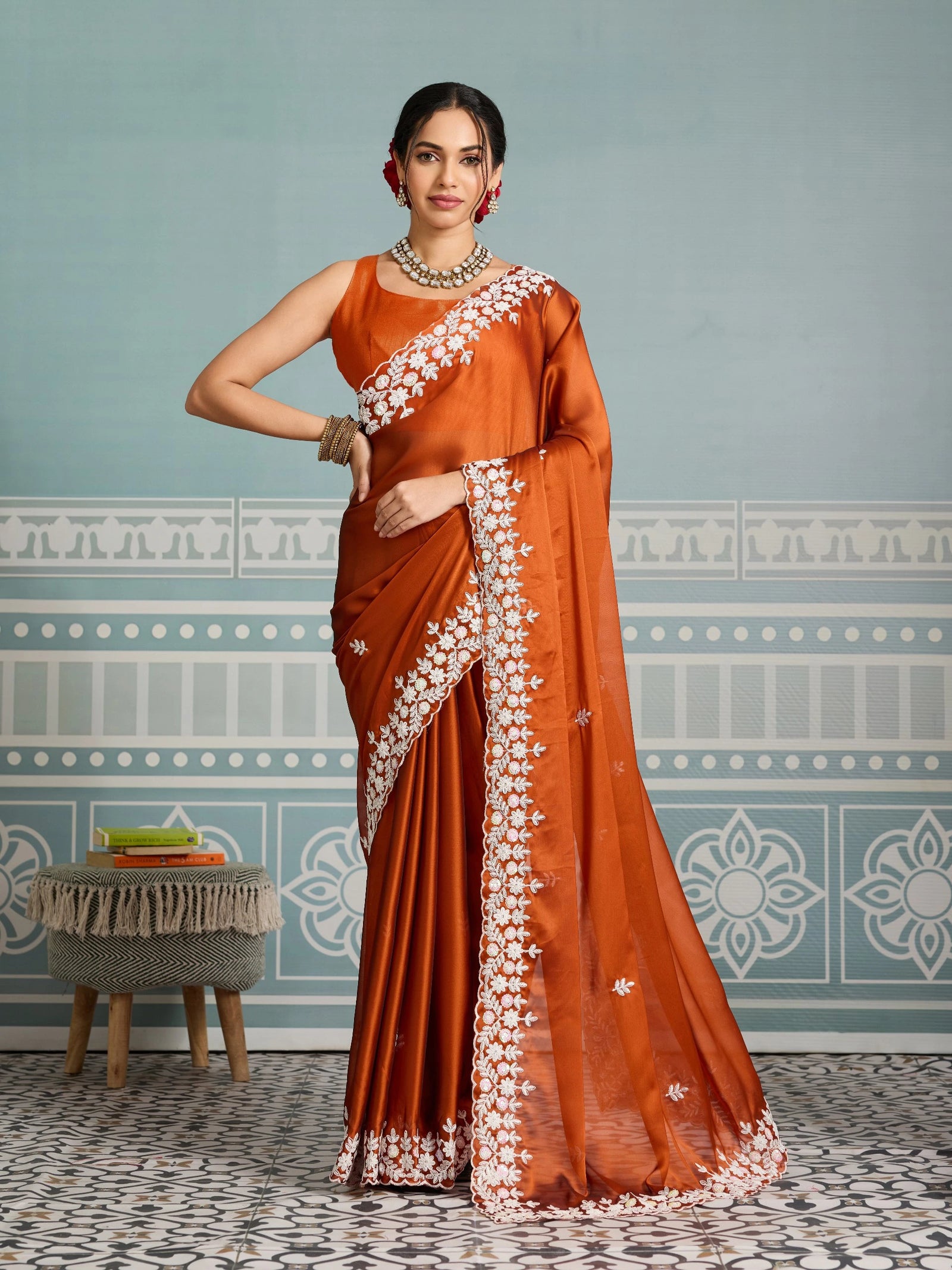 Woman wearing a traditional orange saree with white patterns against a decorative wall.