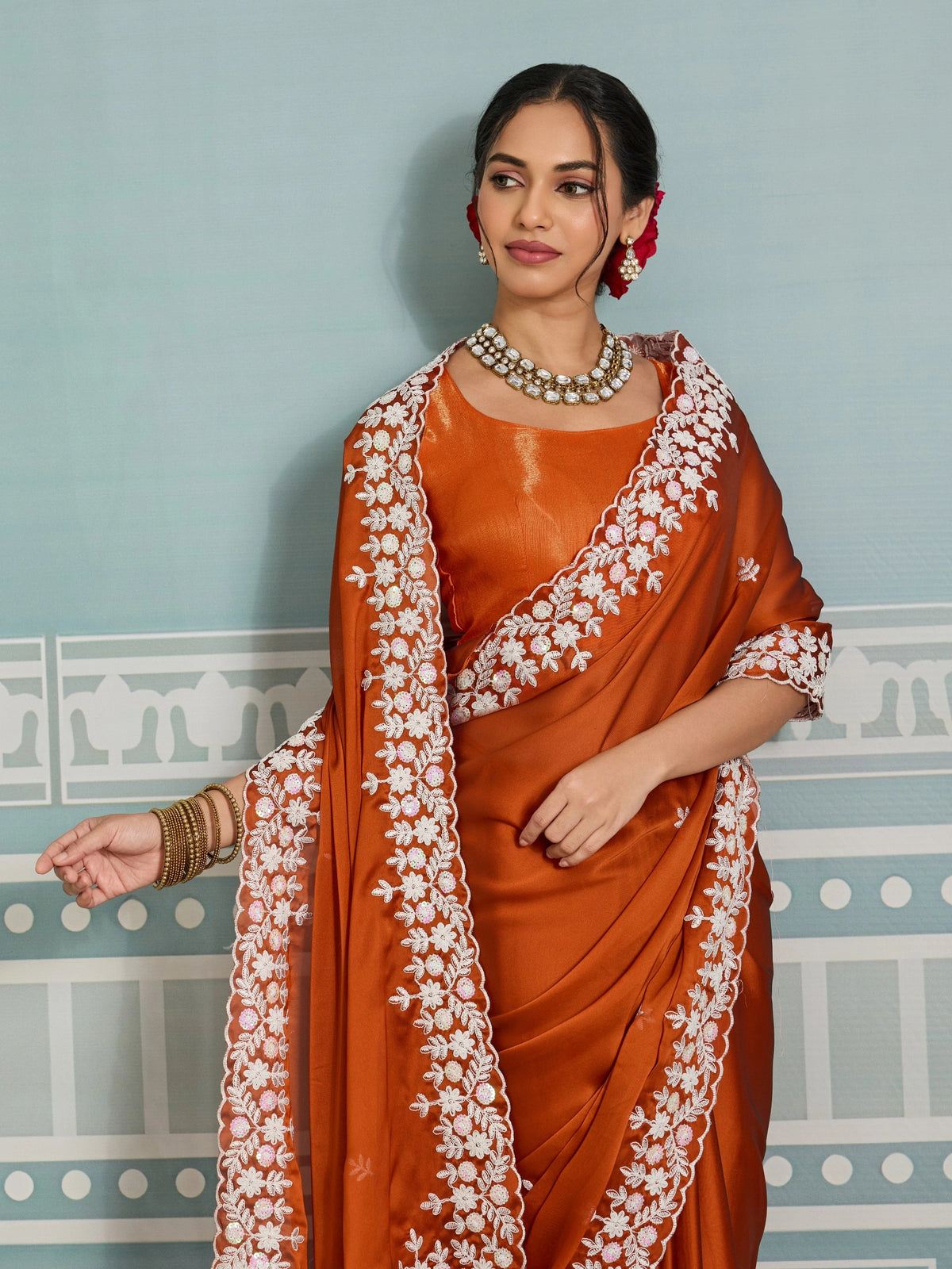 Woman wearing an orange saree with white embroidery against a light blue wall.