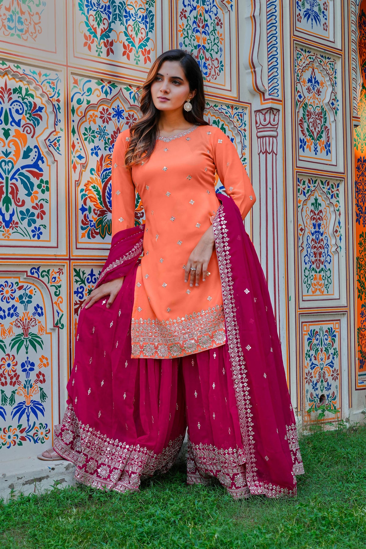 Woman in traditional orange and pink outfit standing in front of a colorful wall.