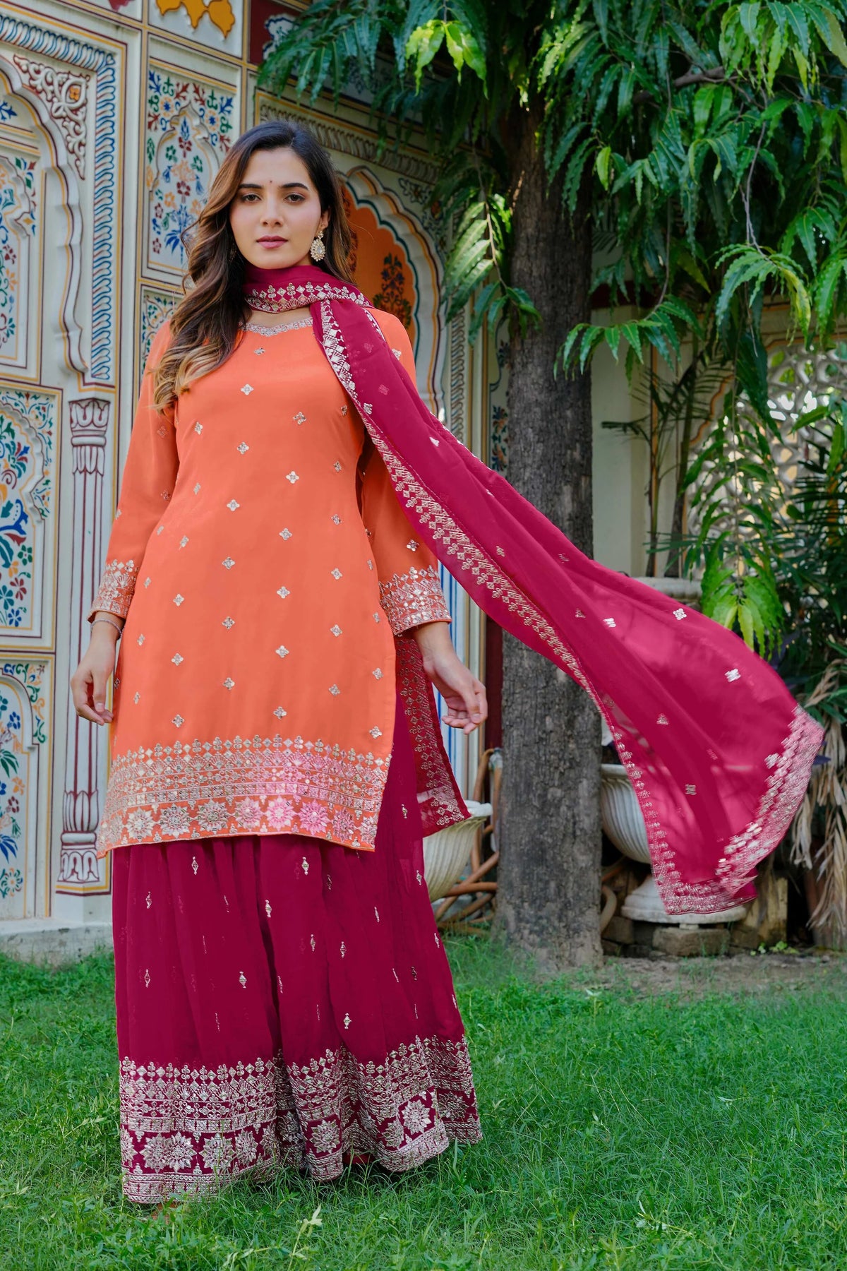 Woman in traditional orange and pink outfit with embroidered patterns standing outdoors.