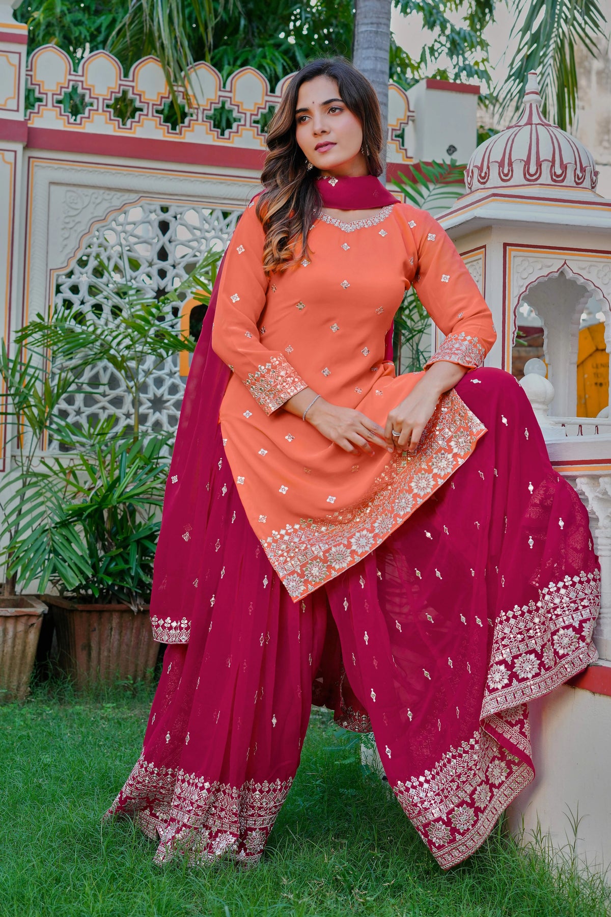 Woman in traditional pink and orange outfit standing outdoors with decorative wall and plants in the background