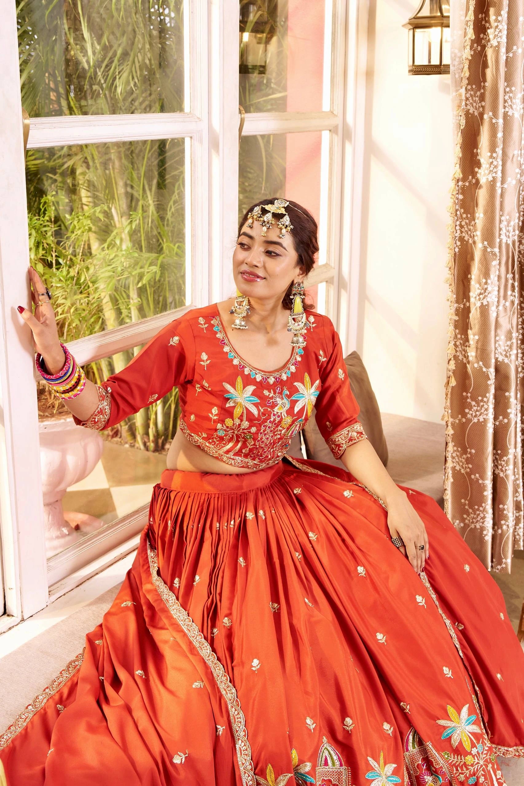 Woman in a red traditional outfit with floral patterns sitting by a window.