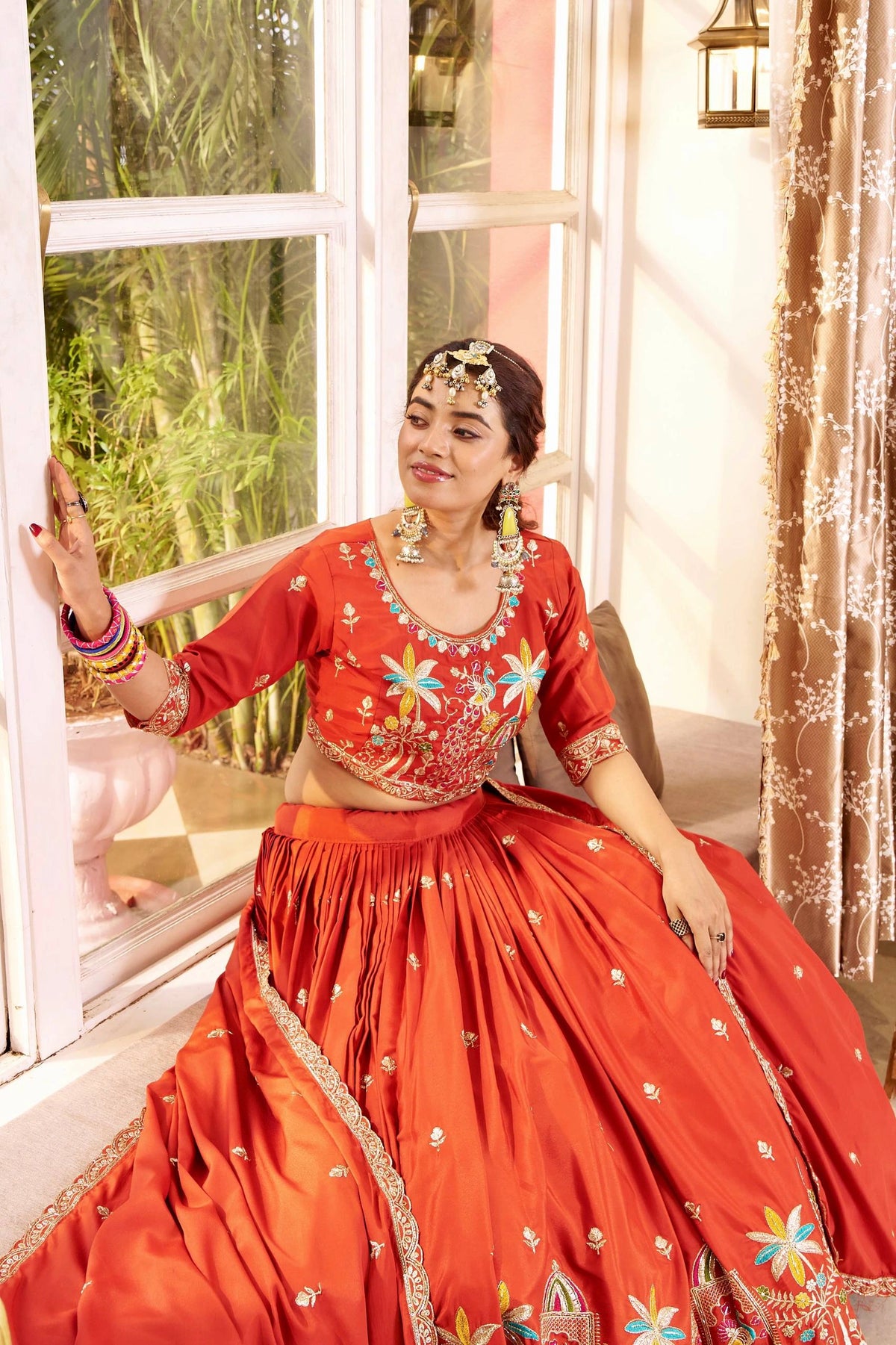 Woman in a red traditional outfit with floral patterns sitting by a window.