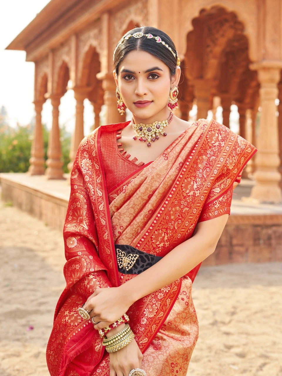 Woman in a red saree with gold patterns standing in front of an architectural background