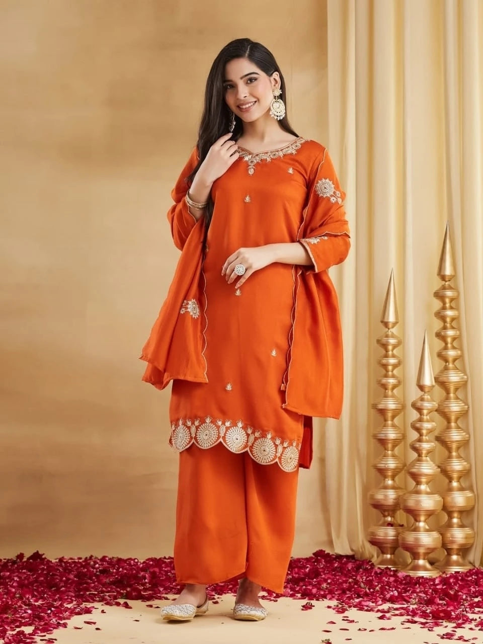 A full-length studio portrait of a woman wearing a rust-orange raw silk kurti set. The short kurti features detailed gold zari work on the V-neck and sleeves, with a distinctive gold embroidered and scalloped border on the hem. She has a matching dupatta draped over her shoulders. She is standing, smiling, with one hand at her neck, displaying a large ring and matching bangles. She is wearing traditional embellished juttis on a floor covered in red rose petals