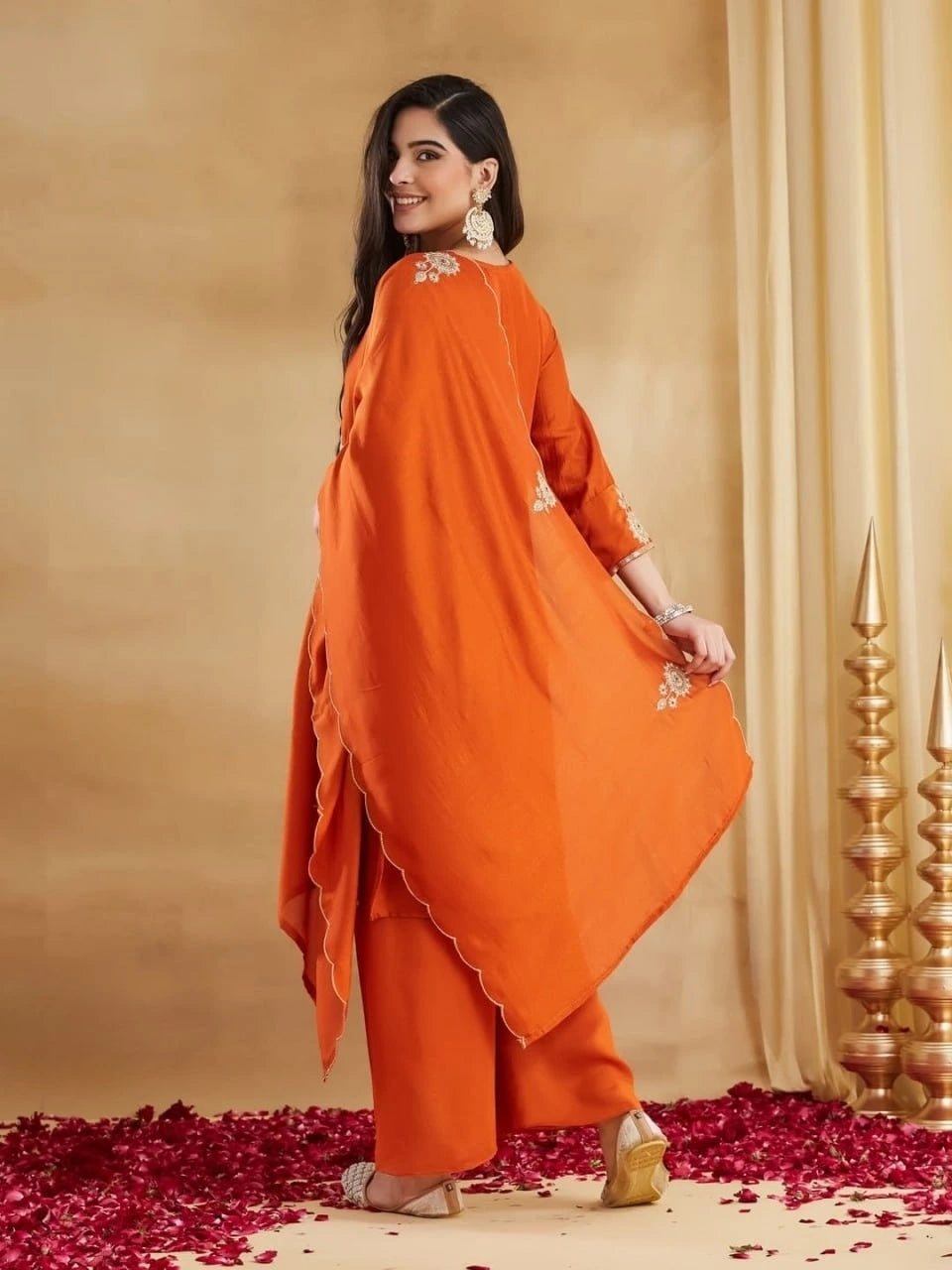 A full-length back view of a woman posing against a light beige textured background with gold drapes. She is wearing a rust-orange ethnic set, holding the edge of her matching scallop-edged dupatta with detailed gold thread embroidery on the corners. The plain wide-legged pants and the kurti sleeve are partially visible. She is wearing embellished beige juttis and has long wavy brown hair.