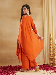 A full-length back view of a woman posing against a light beige textured background with gold drapes. She is wearing a rust-orange ethnic set, holding the edge of her matching scallop-edged dupatta with detailed gold thread embroidery on the corners. The plain wide-legged pants and the kurti sleeve are partially visible. She is wearing embellished beige juttis and has long wavy brown hair.