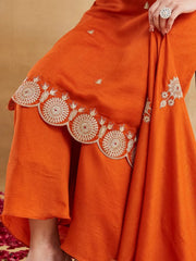 A close-up detail shot of the lower portion of a rust-orange silk kurti set, highlighting the intricate embroidery. The heavy, gold-thread embroidery features traditional circular motifs and patterns along the scalloped hemline of the kurta. A matching dupatta is draped, showing similar gold threadwork. A partial view of a woman's leg wearing a large cocktail ring on her finger is included, along with red rose petals on the floor in the foreground.