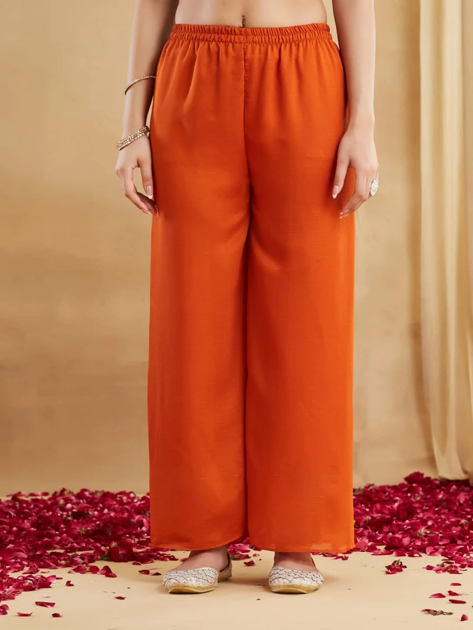 Person wearing orange pants standing on a beige background with red flower petals.