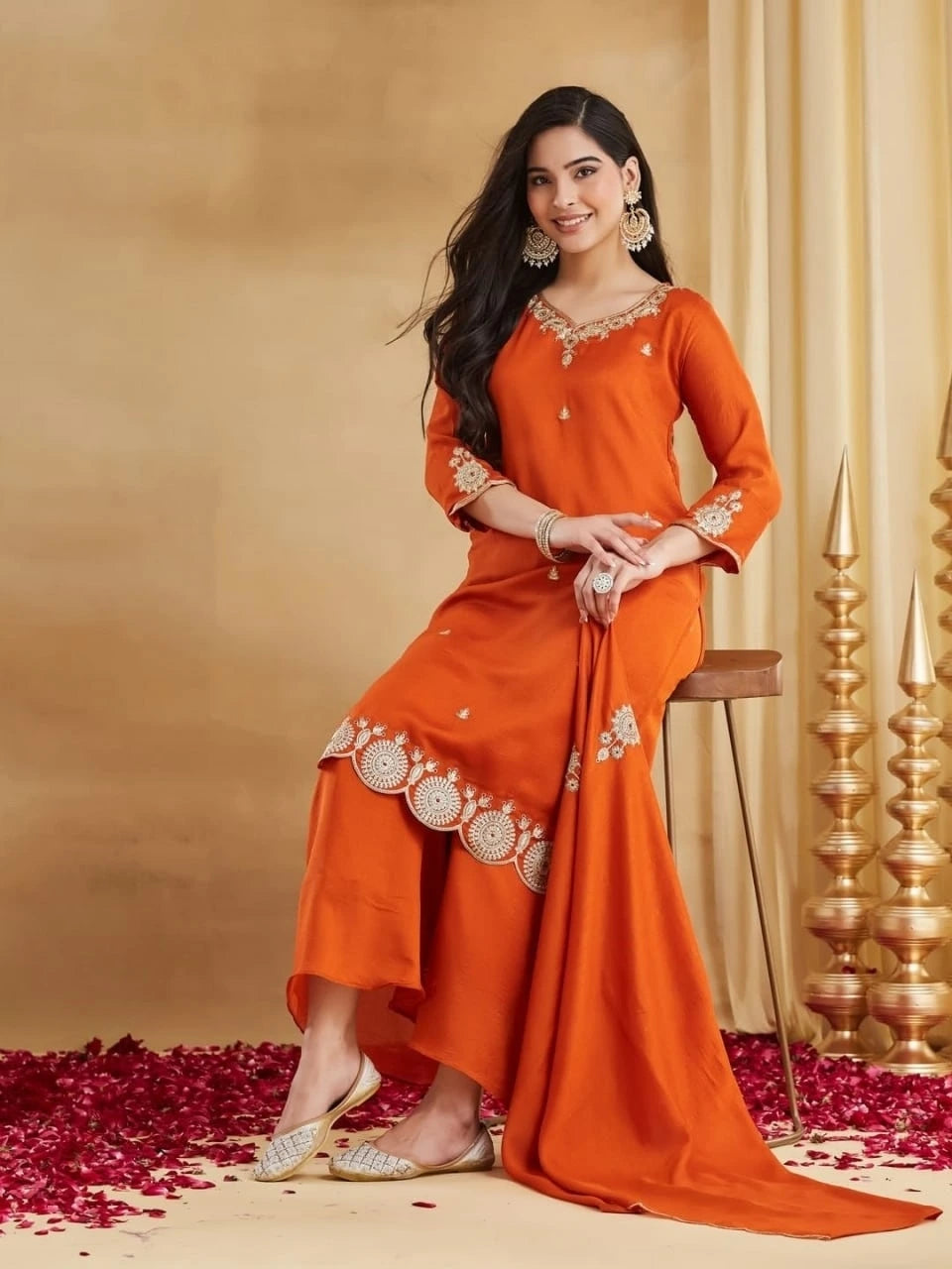 A woman is seated gracefully on a wooden stool, smiling and looking toward the camera. She is wearing a rust-orange Raw Silk Kurti set. The short kurta features intricate gold thread embroidery on the neck, sleeves, and a prominent scalloped embroidered hemline. She has wide-legged trousers and matching juttis. Her look is accessorized with heavy gold traditional earrings, bangles, and a cocktail ring. The floor is scattered with red rose petals, and gold pyramid structures are visible in the background