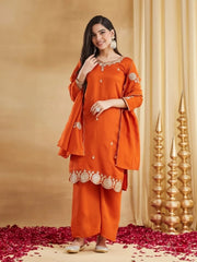 A full-length studio portrait of a woman wearing a rust-orange raw silk kurti set. The short kurti features detailed gold zari work on the V-neck and sleeves, with a distinctive gold embroidered and scalloped border on the hem. She has a matching dupatta draped over her shoulders. She is standing, smiling, with one hand at her neck, displaying a large ring and matching bangles. She is wearing traditional embellished juttis on a floor covered in red rose petals
