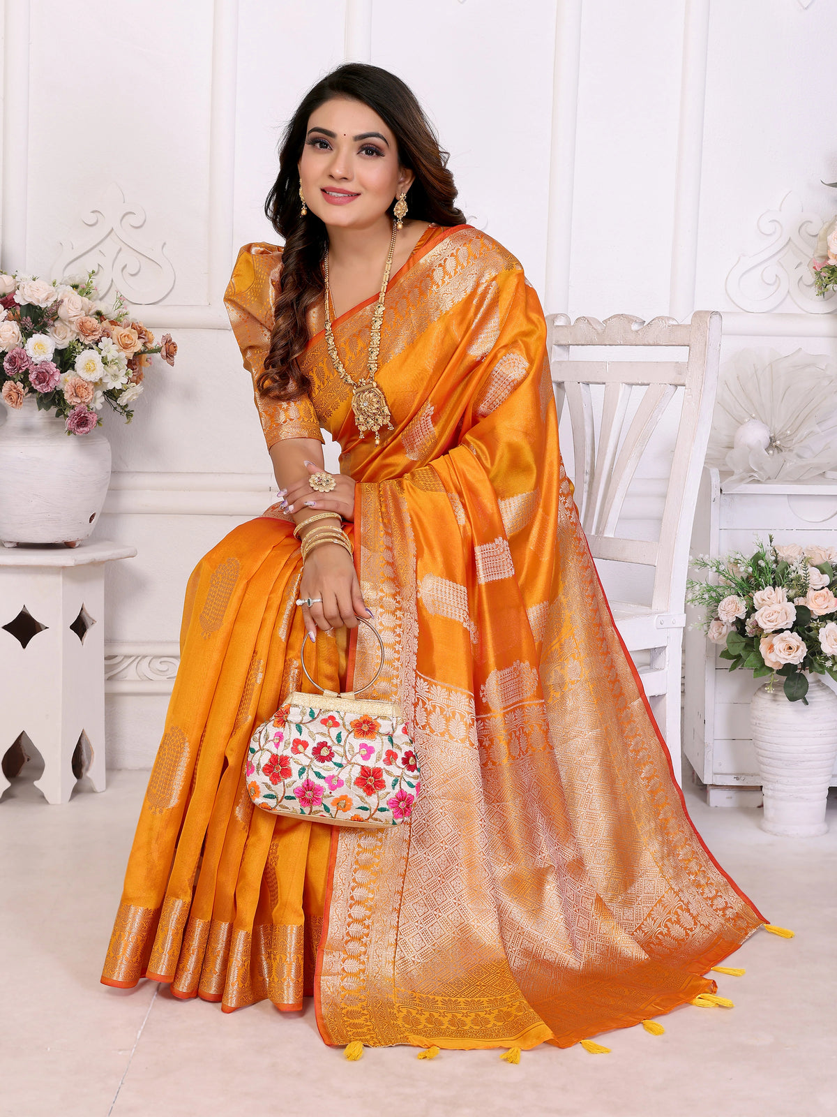 Woman in an orange saree with a floral handbag in a decorated room.