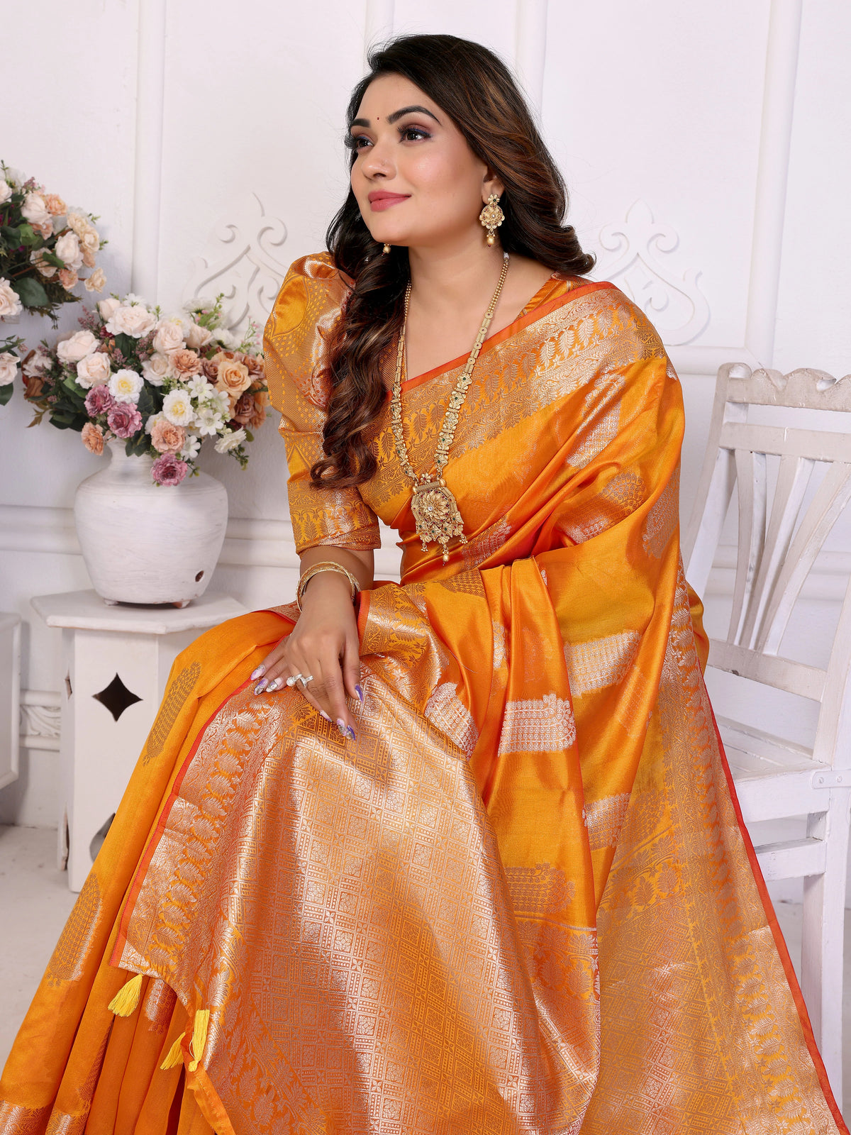 Woman in an orange saree with gold patterns sitting on a white chair.