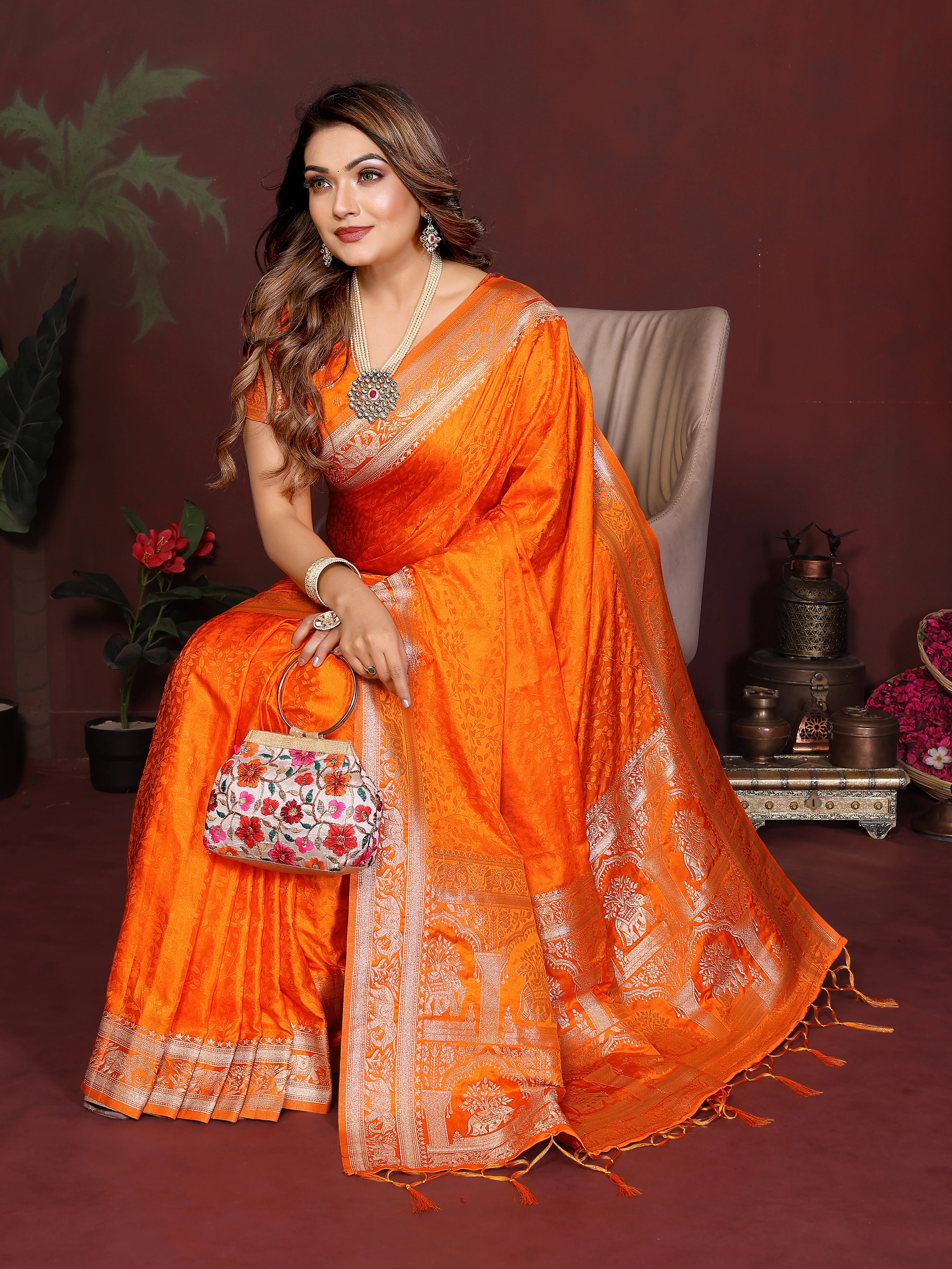 Woman in an orange saree with a decorative bag, sitting against a dark background.