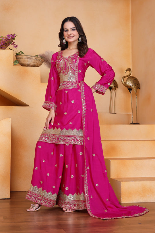 Woman in a bright pink traditional outfit with gold embroidery standing indoors.