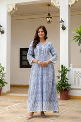 Woman in a blue patterned dress standing in an elegant interior setting with plants and decorative lights.