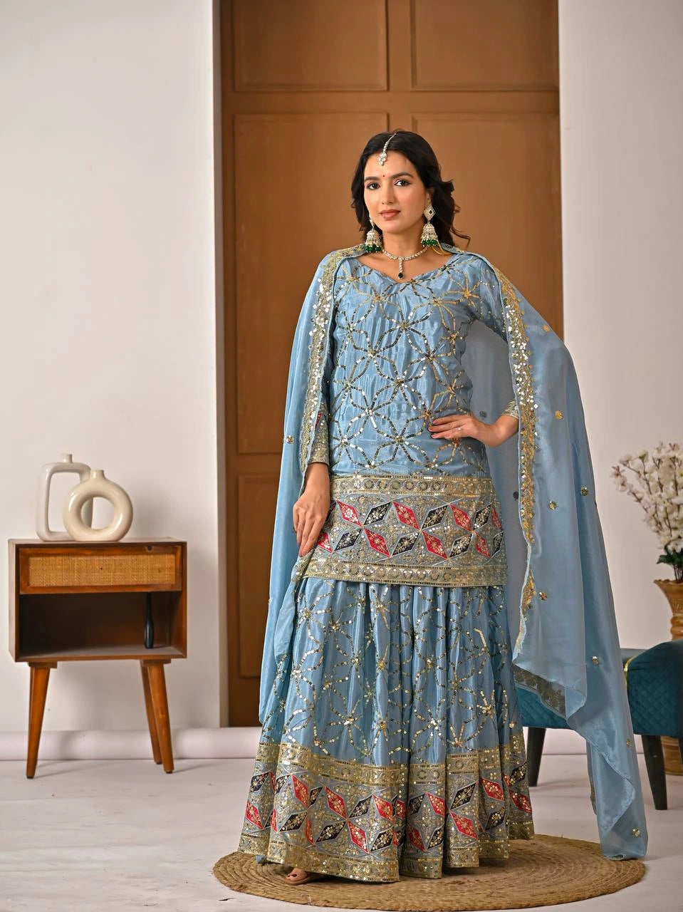 Woman wearing a light blue embroidered traditional outfit in a room with wooden furniture.