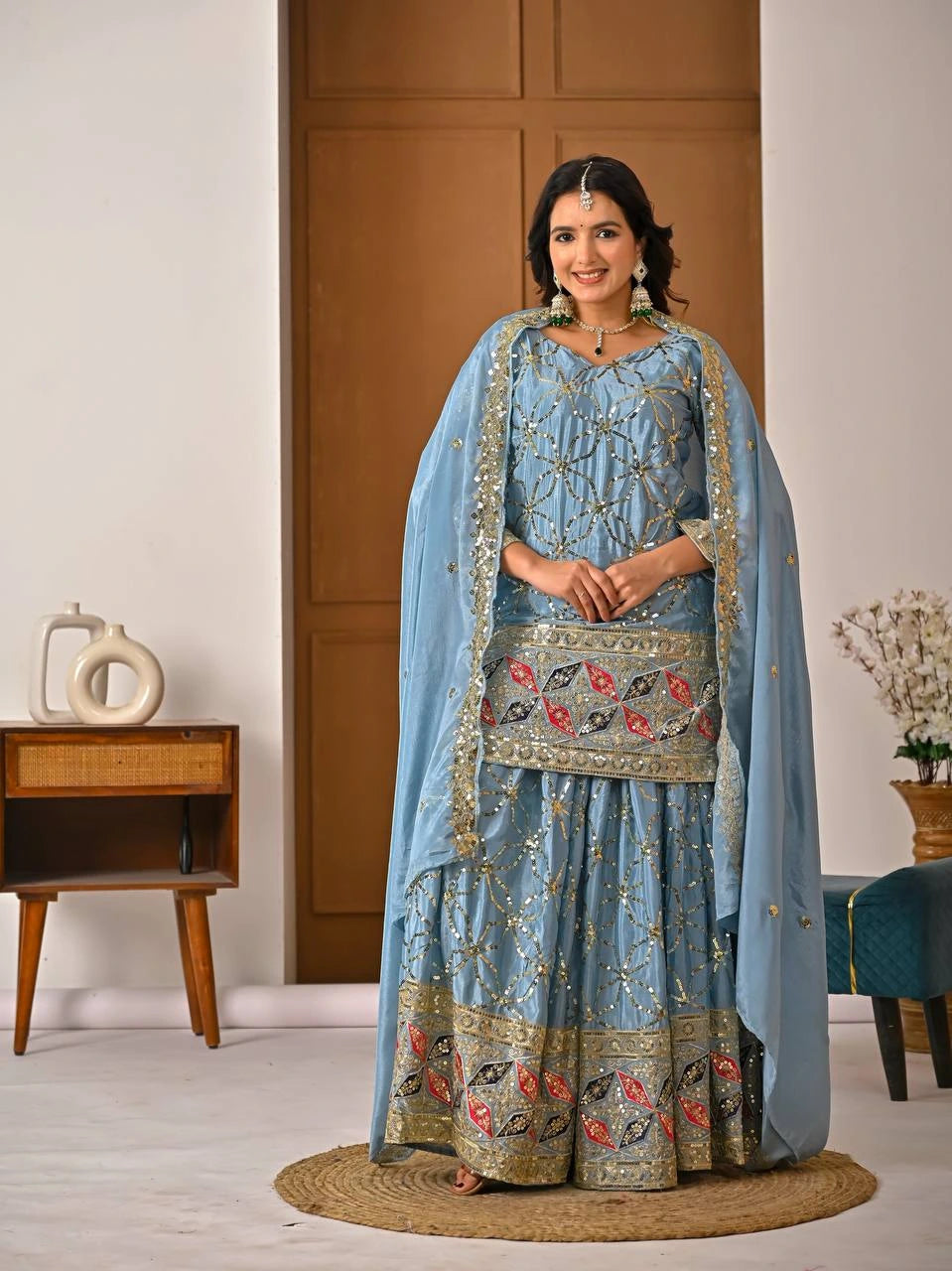 Woman in a traditional light blue embroidered outfit standing in a room with wooden furniture.