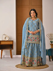 Woman in a traditional light blue embroidered outfit standing in a room with wooden furniture.