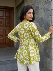 A rear-view shot of a woman looking back over her shoulder, showcasing the gathered waist and floral pattern of her green tunic paired with white pants.
