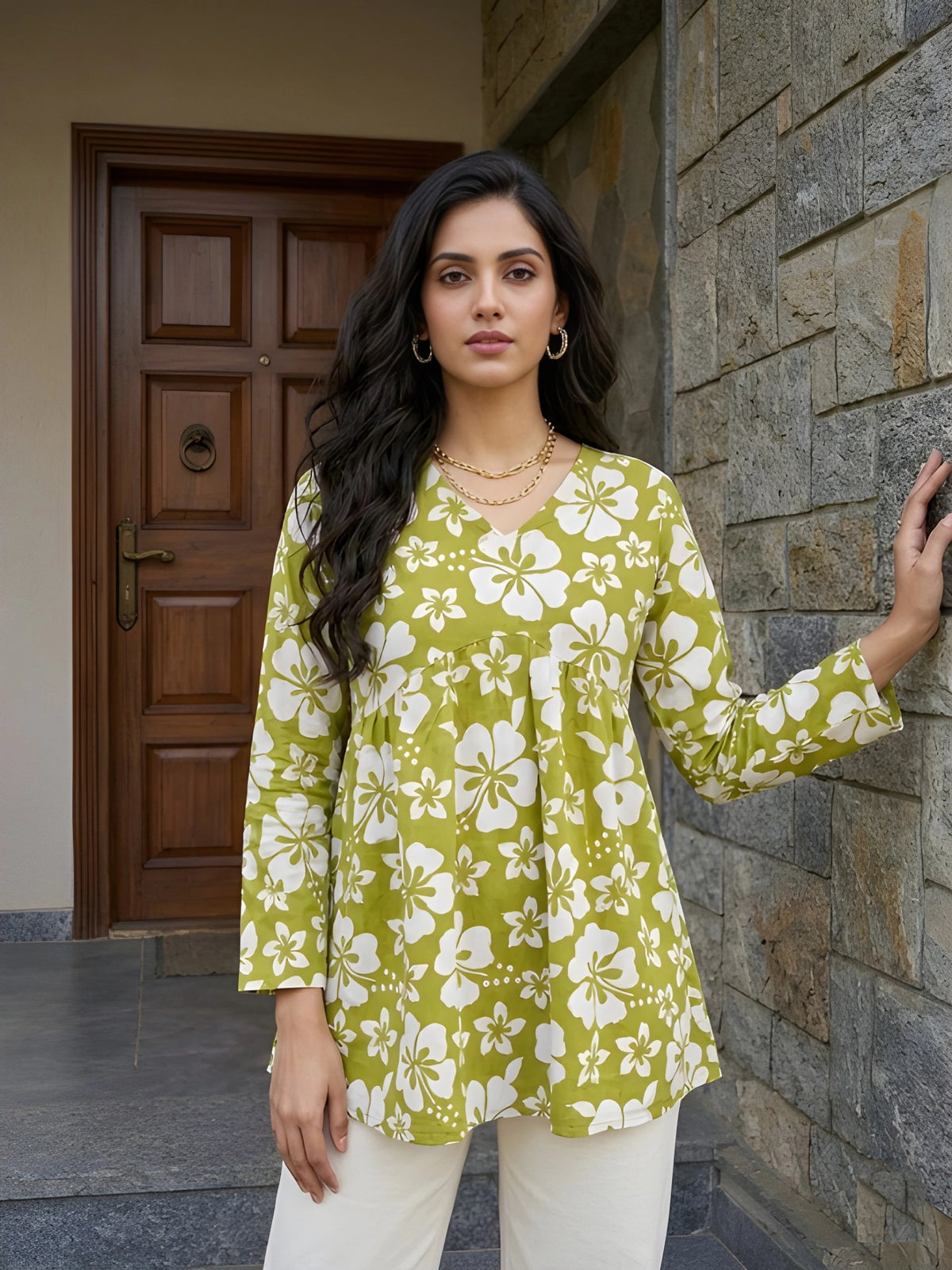 A medium shot of a woman posing in a green floral tunic and white pants, resting one hand against a textured stone wall.