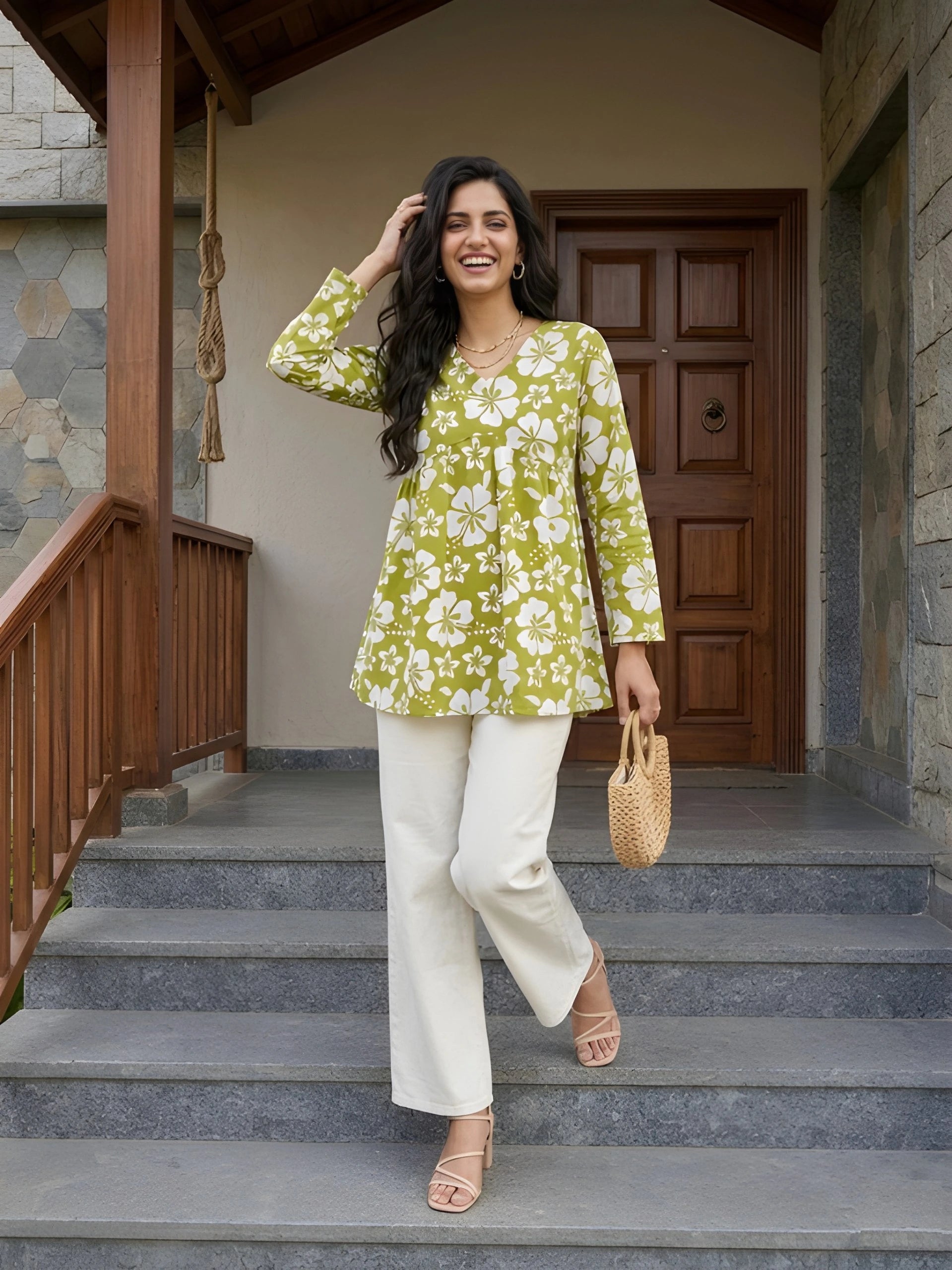 A full-length shot of a woman laughing and walking down stone steps, carrying a small woven handbag and wearing a green floral tunic with white pants.