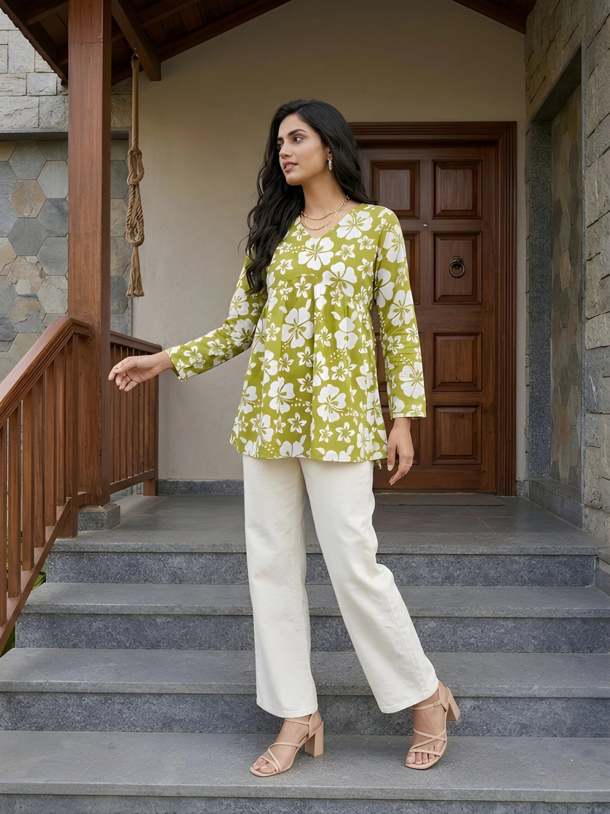 A full-length shot of a woman standing on stone steps, wearing a green and white floral V-neck tunic with white trousers, looking off to the side.